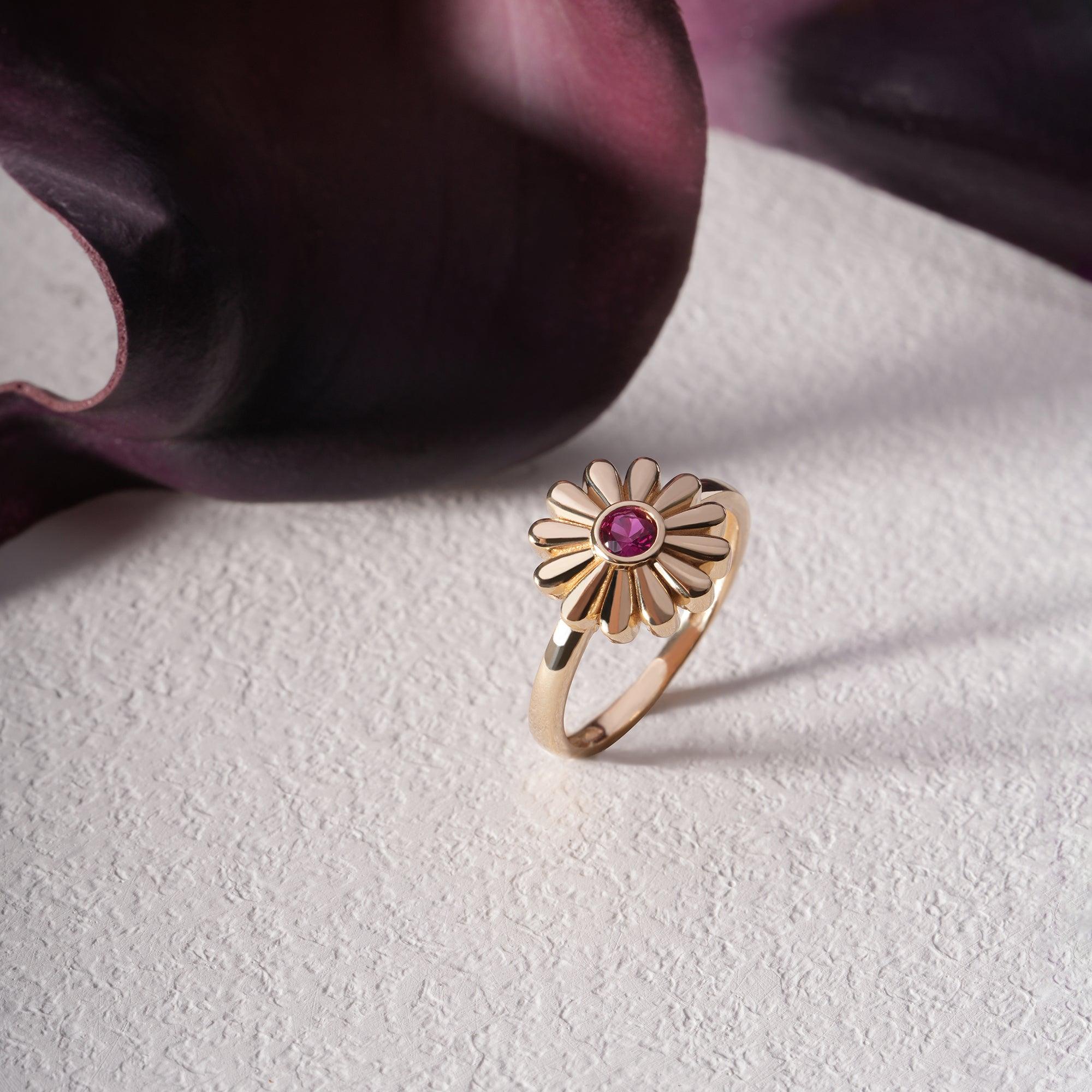 Gold flower ring with pink gemstone center on textured white surface, elegant jewelry