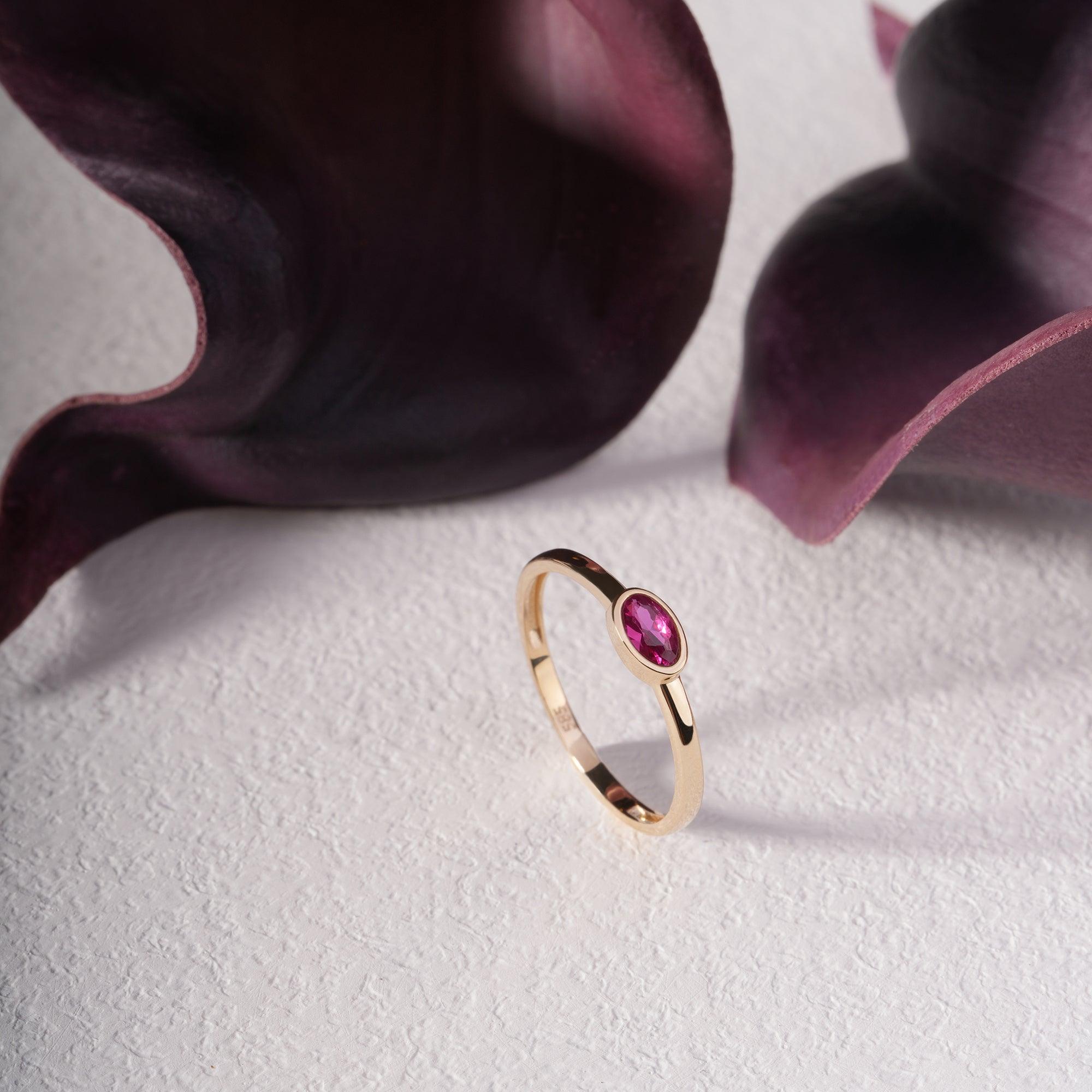 Gold ring with oval pink gemstone on white textured surface, styled with deep purple petals