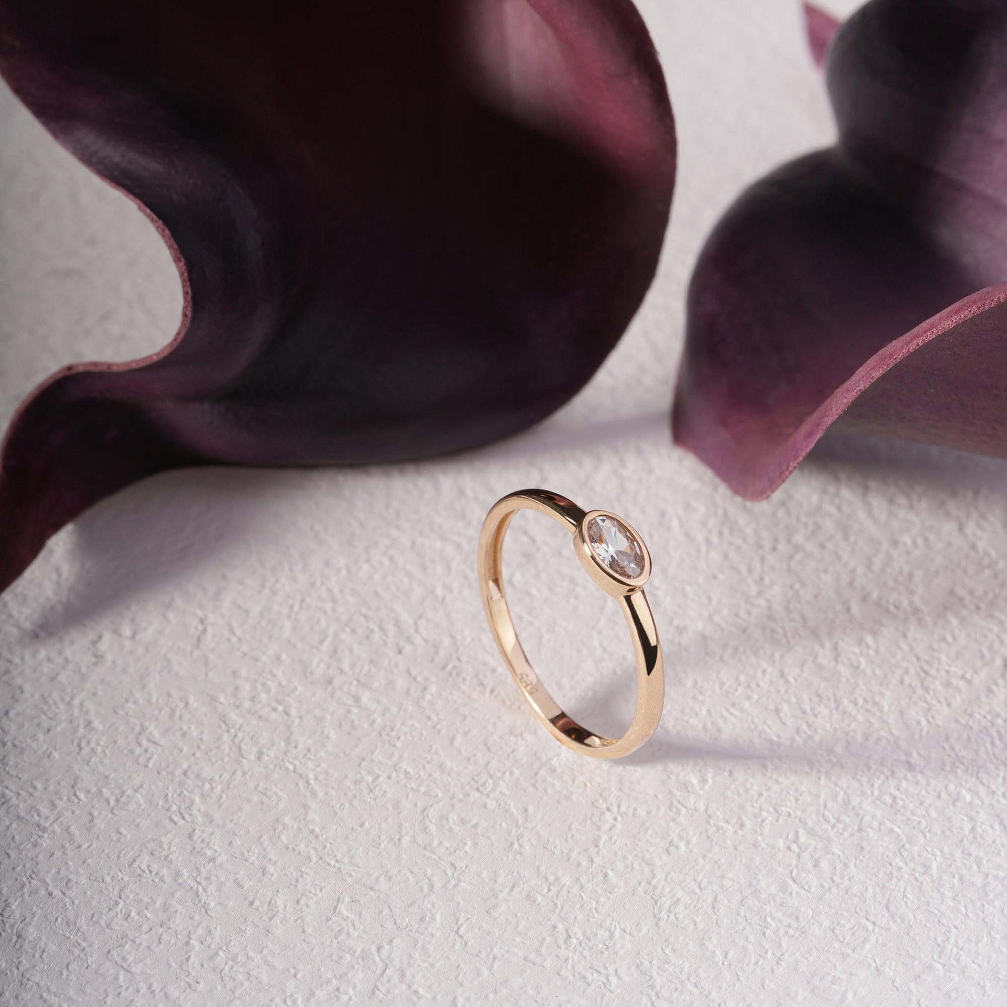 Elegant gold ring with clear gemstone showcased on textured surface with purple flowers