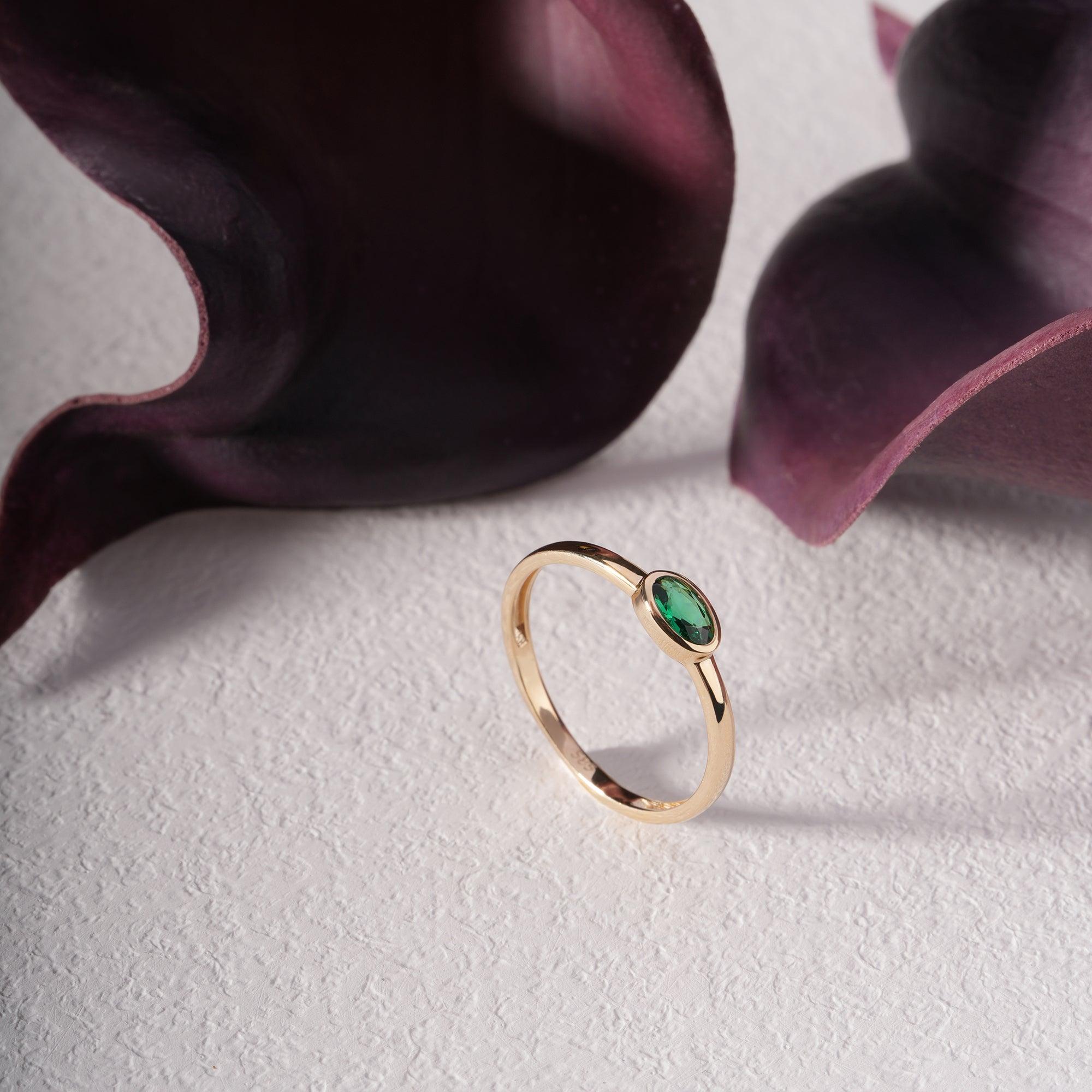 Gold ring with oval green gemstone on textured white surface, purple calla lilies nearby