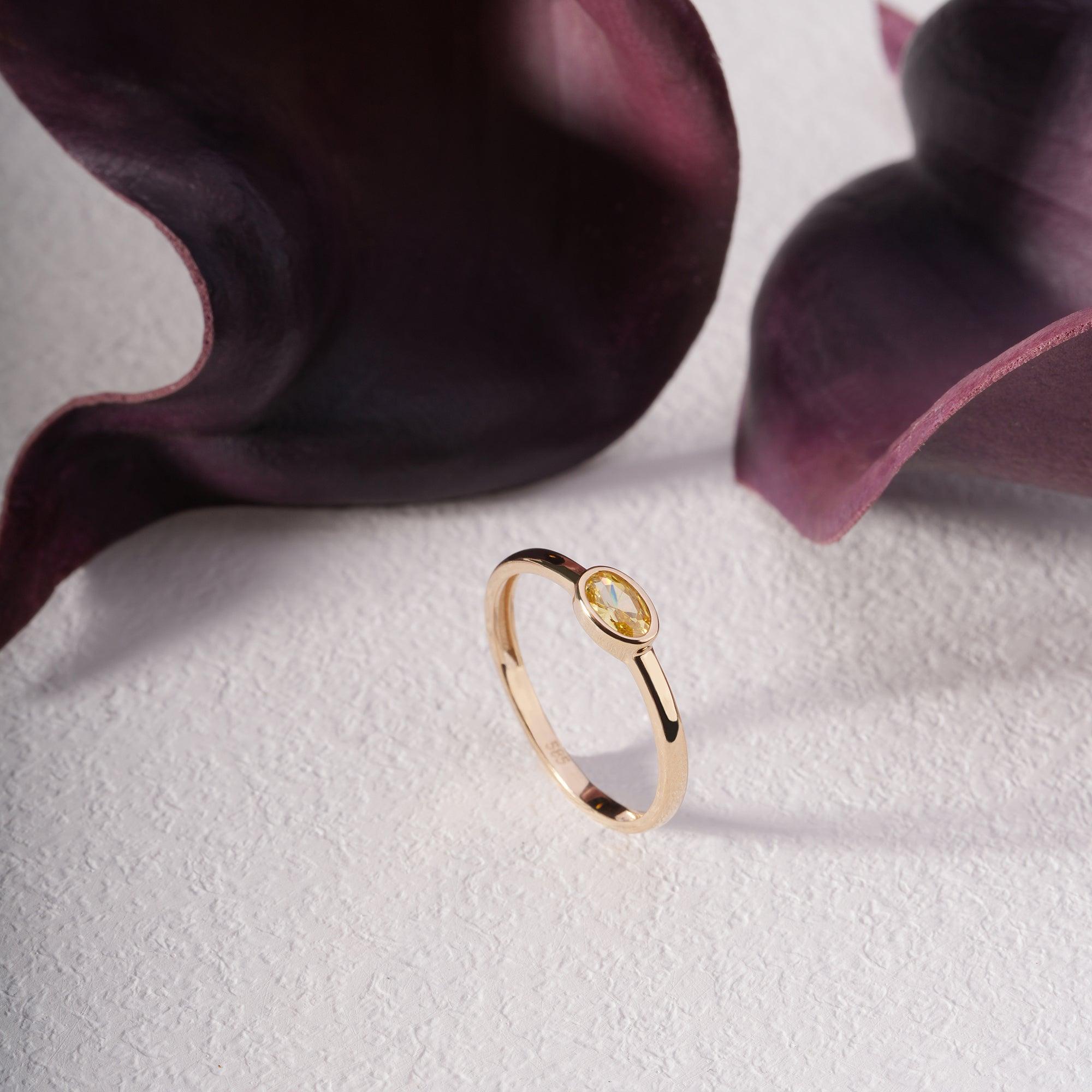 Elegant gold ring with oval yellow gemstone on textured white surface, floral backdrop