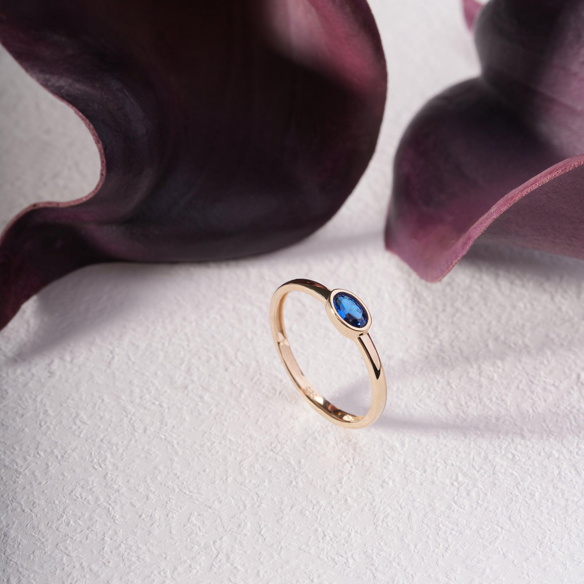 Gold ring with oval blue gemstone on textured white surface, accented by dark purple leaves