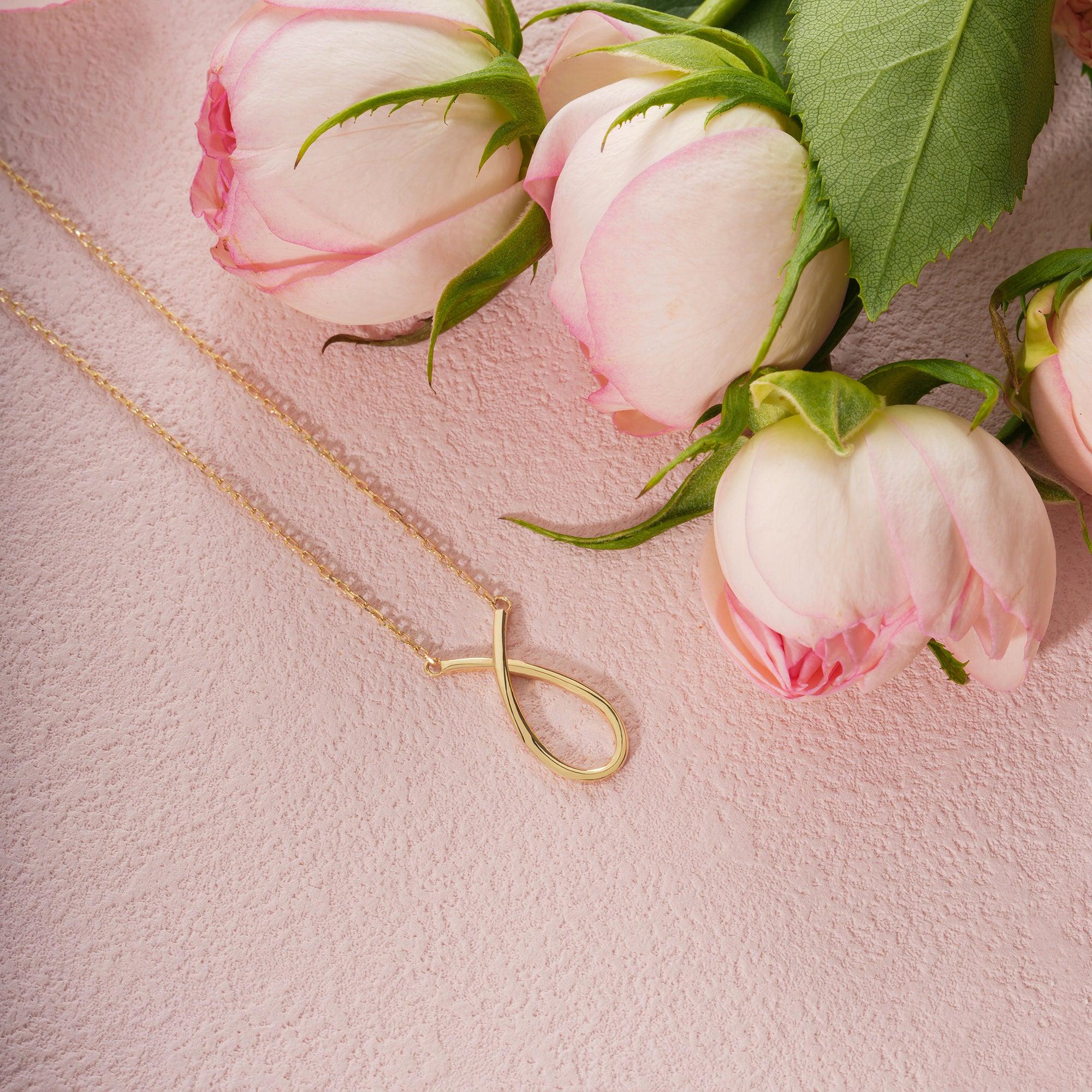 Gold infinity necklace on pink textured surface with pale pink roses