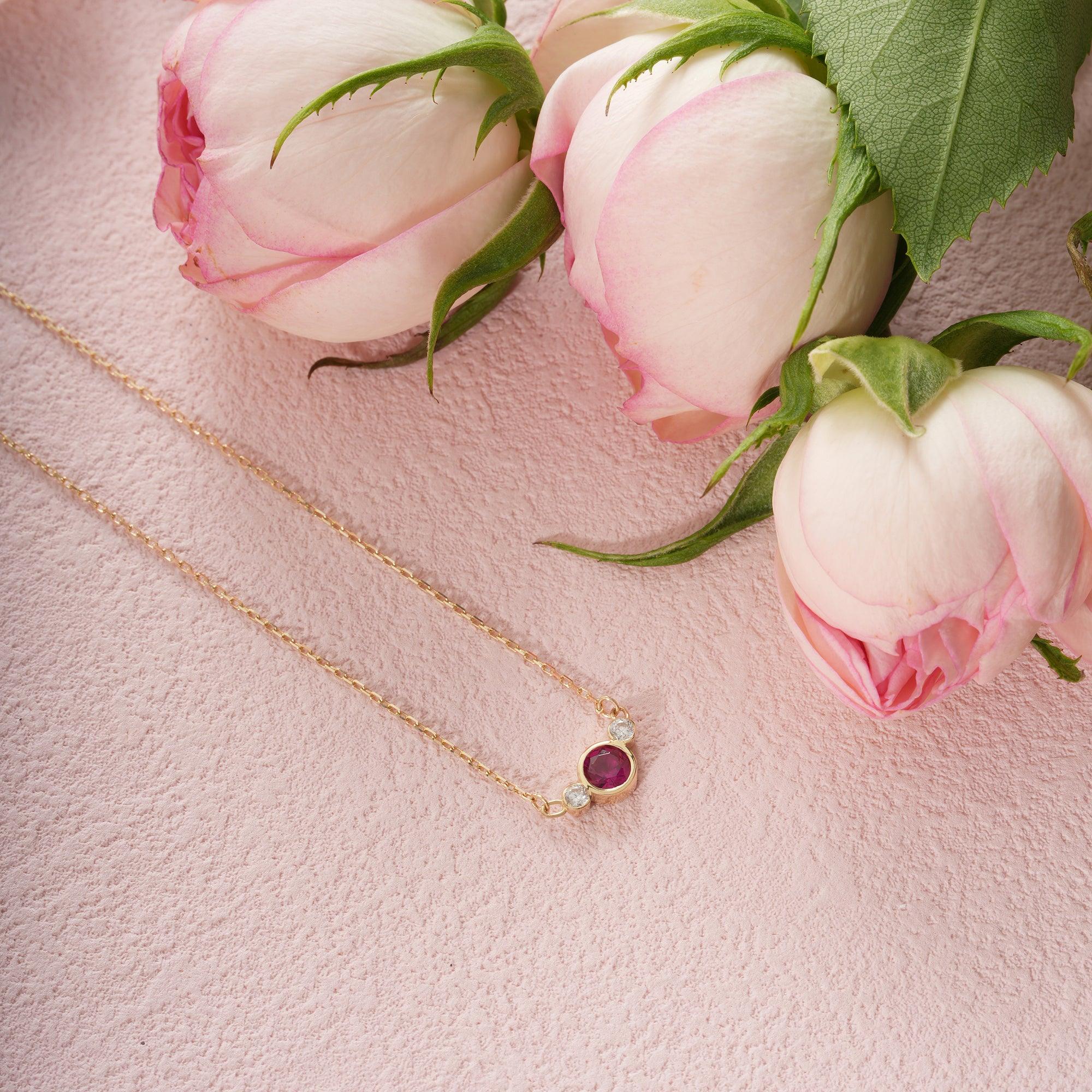 Gold necklace with red gemstone pendant and diamond accents on pink textured surface, surrounded by pink roses
