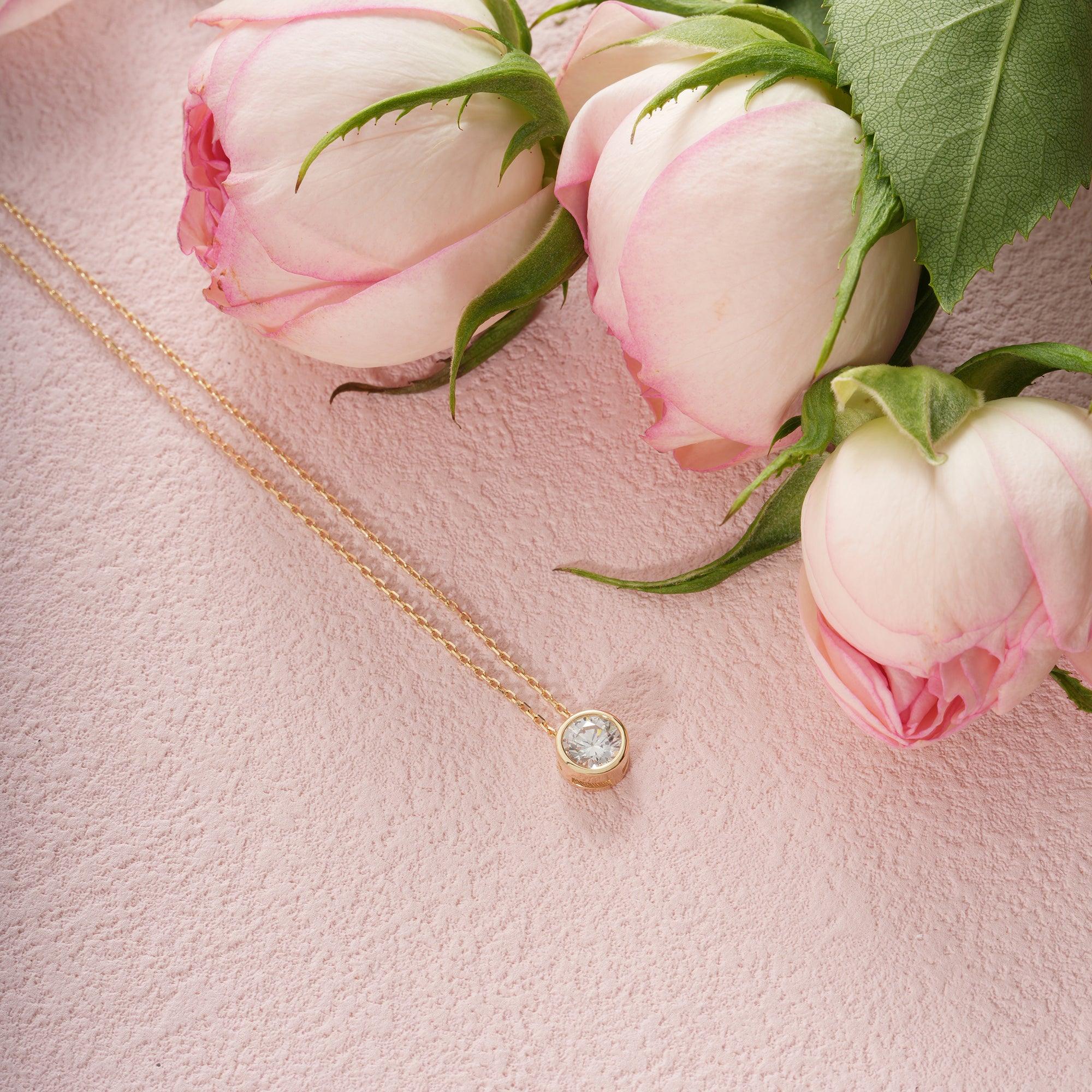 Gold necklace with round diamond pendant on pink textured background near pink roses