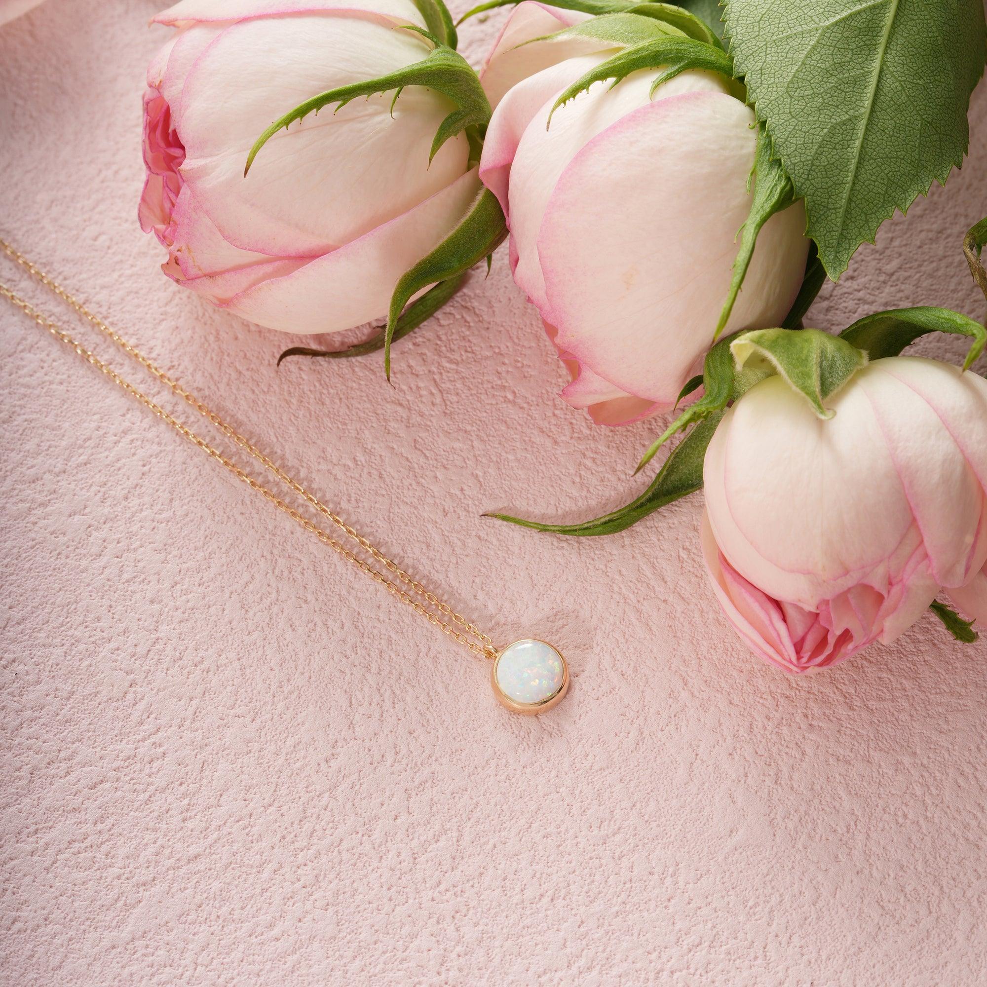 Gold necklace with opal pendant on pink textured surface next to pink rosebuds