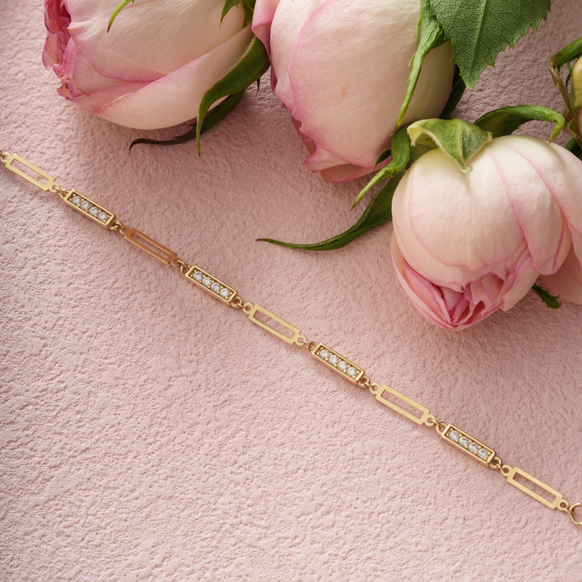 Gold bracelet with rectangular diamond links on pink textured surface beside pink roses