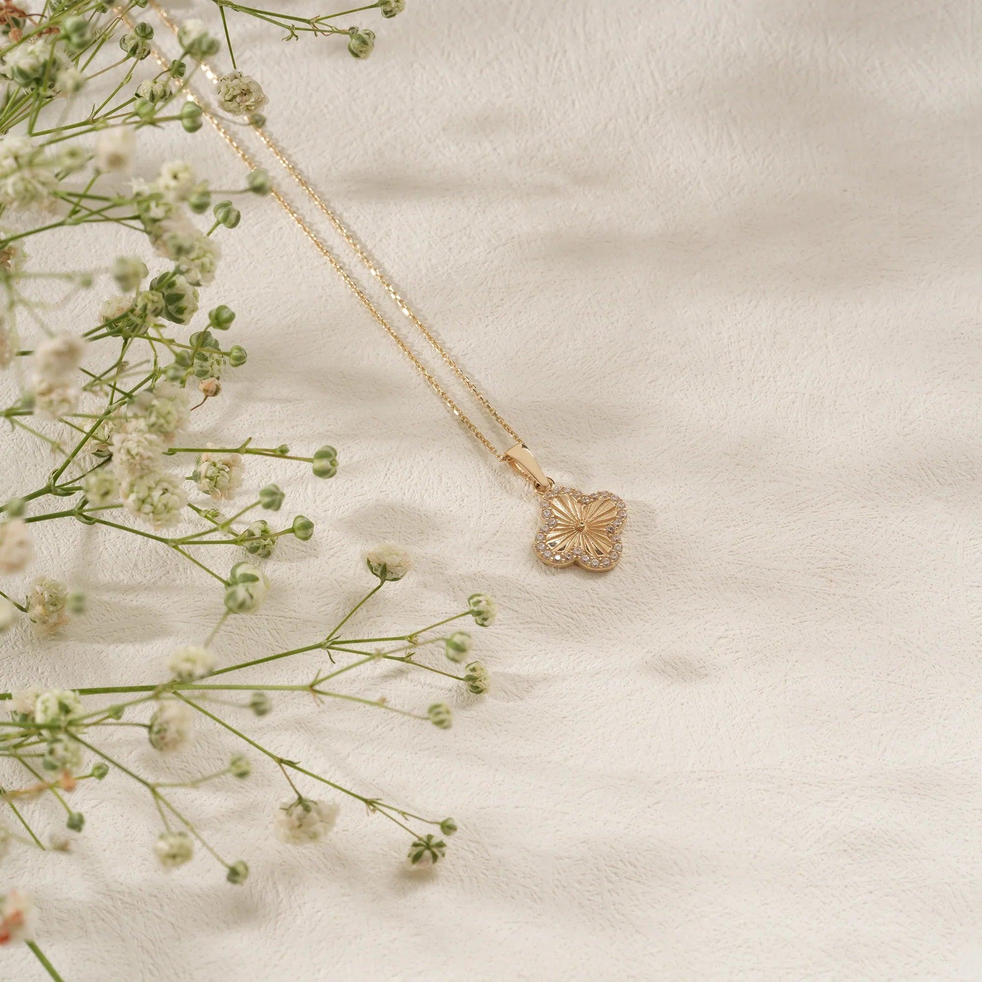 Gold clover pendant necklace on textured ivory background with delicate white flowers
