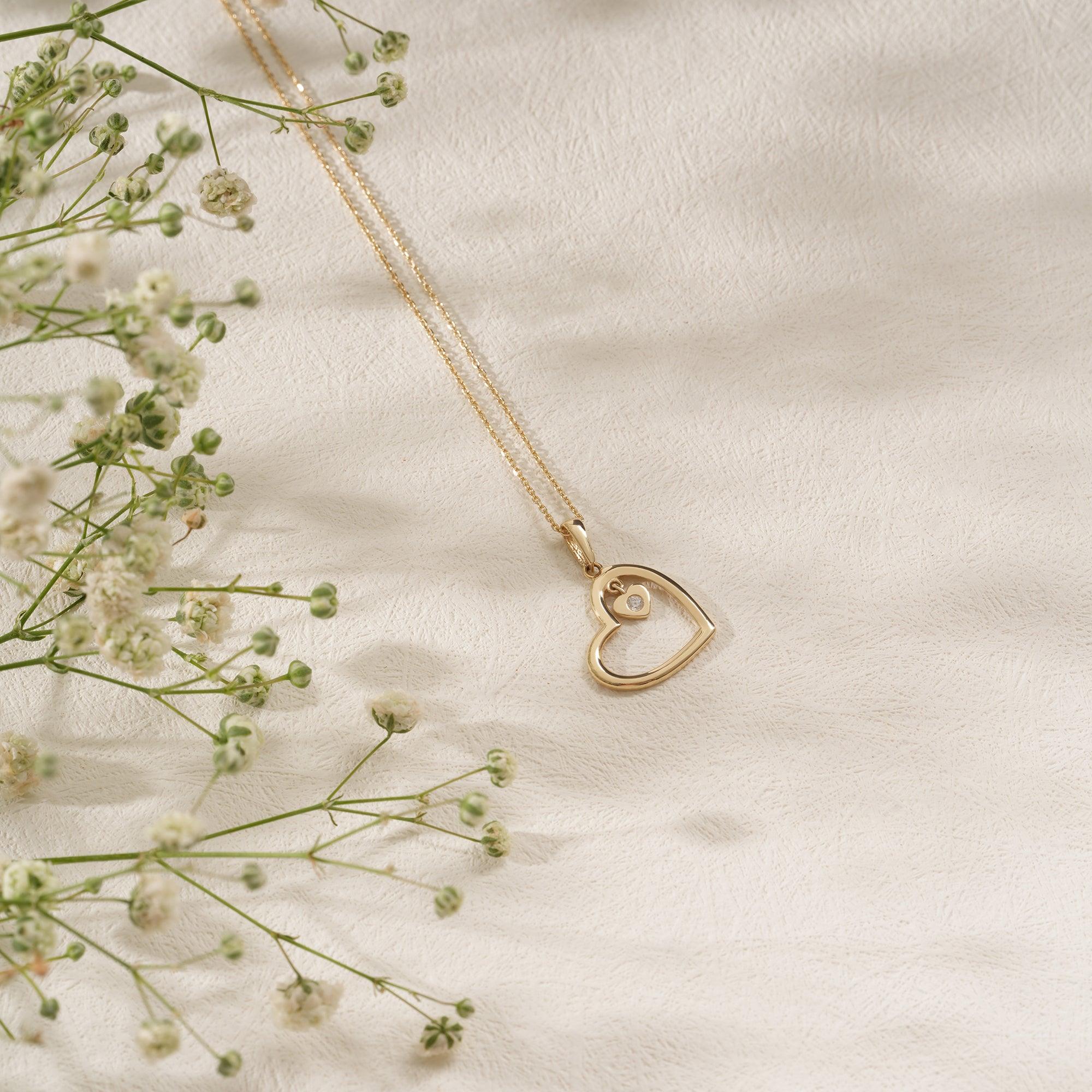 Gold heart pendant necklace with small diamond on textured surface beside white flowers