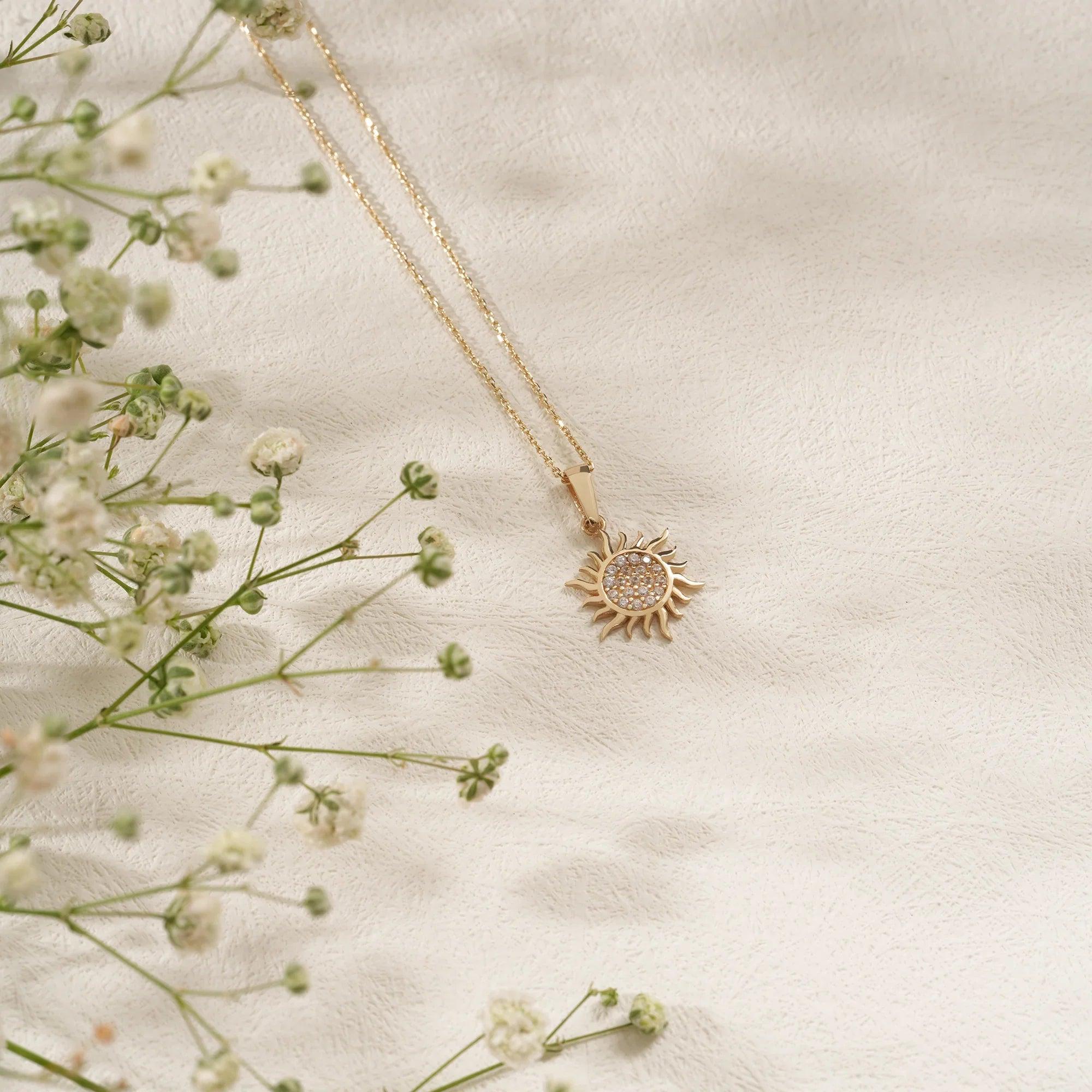 Gold sun pendant necklace with crystal details on a delicate chain, displayed on fabric with small white flowers.