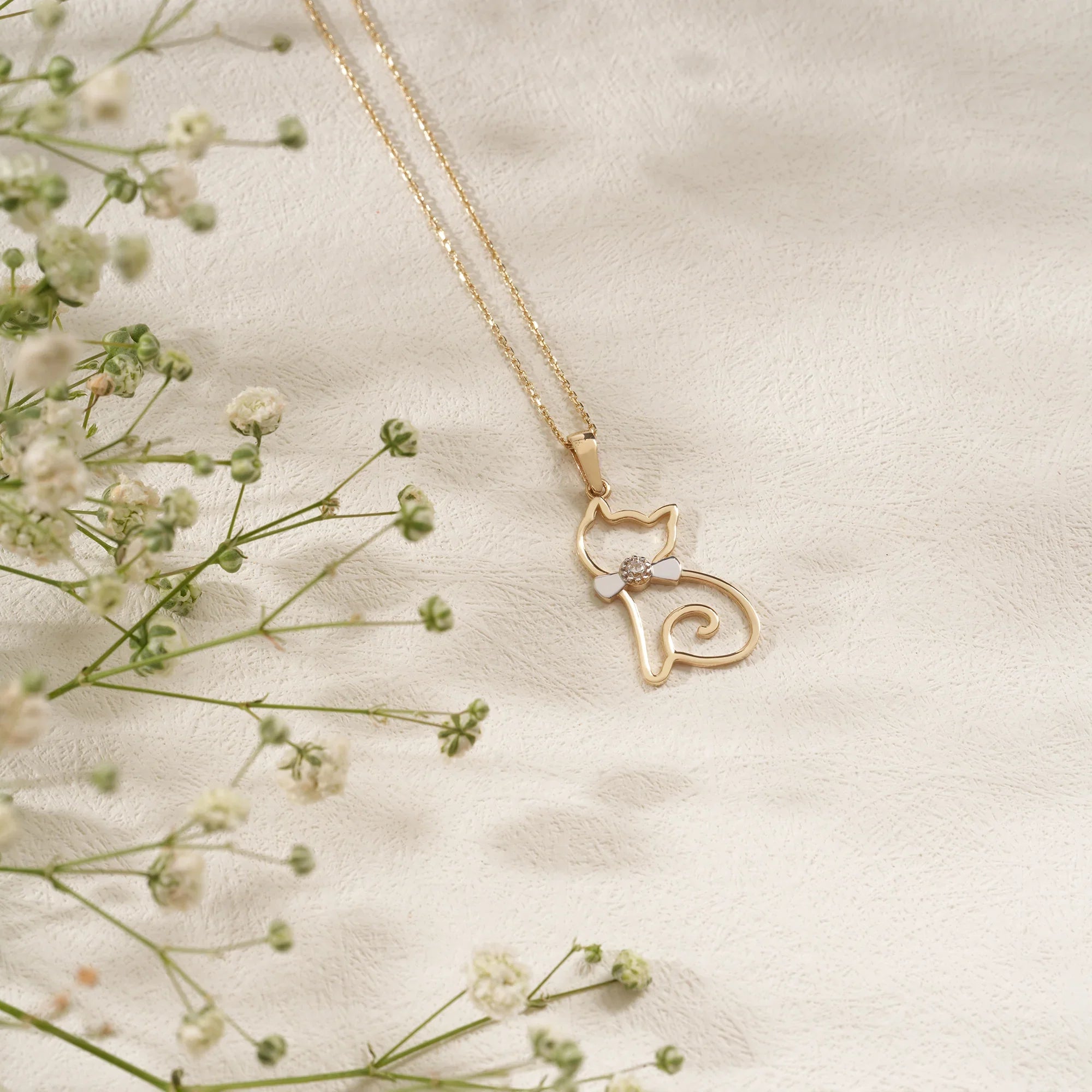 Gold cat-shaped necklace with a small heart pendant on a light fabric background with delicate flowers.