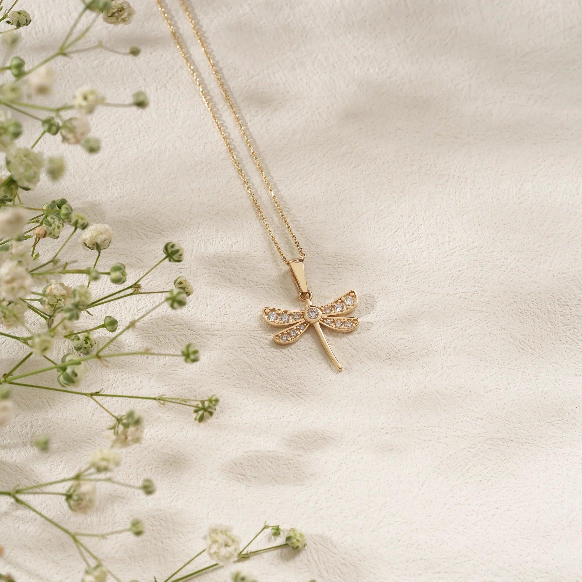 Gold dragonfly pendant necklace with cubic zirconia on textured white background and flowers
