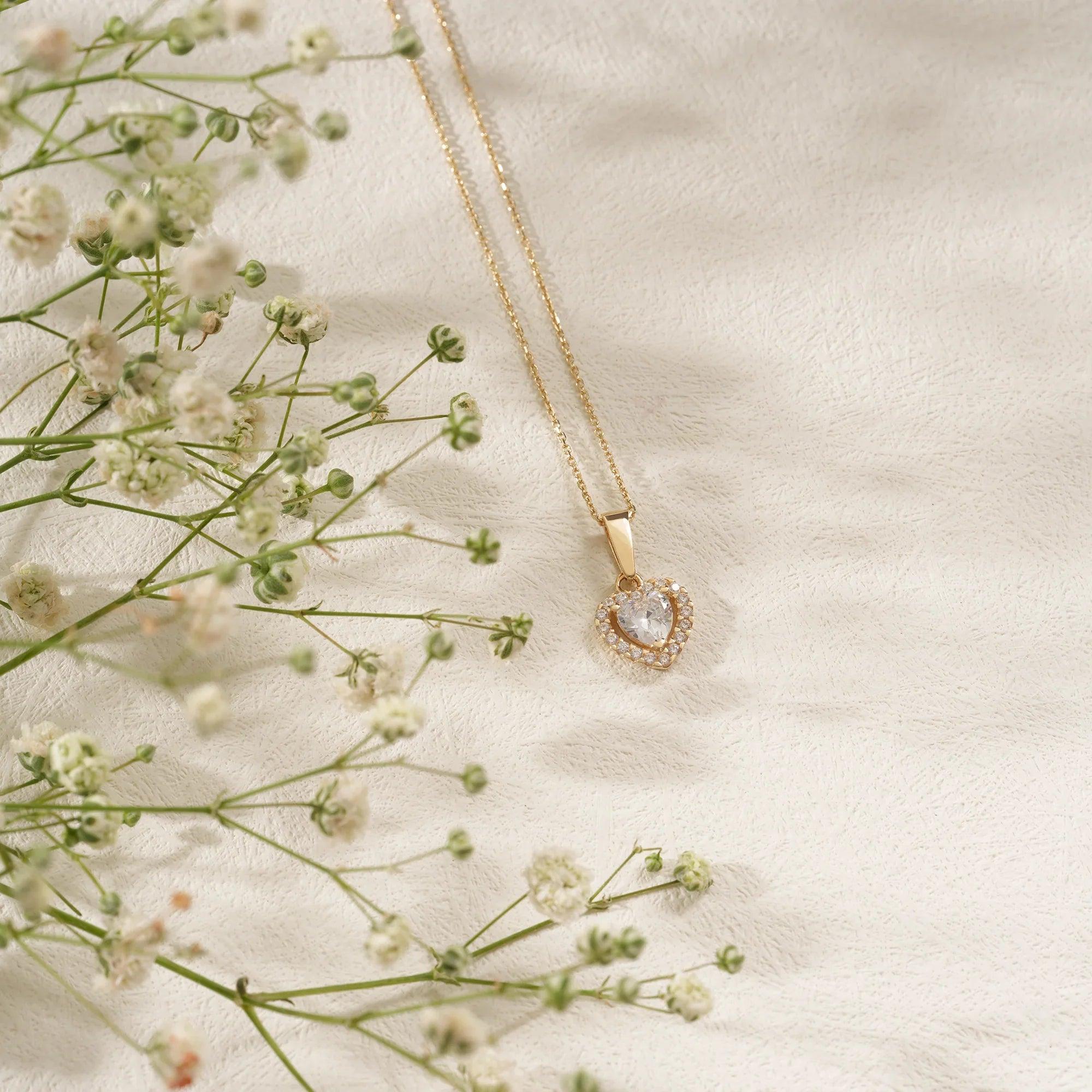 Gold heart-shaped pendant necklace with clear gemstone, styled on soft cream fabric with flowers