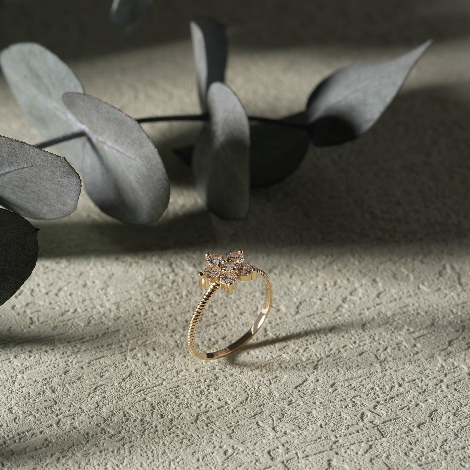 Gold ring with diamond flower design on textured surface, eucalyptus leaves in background
