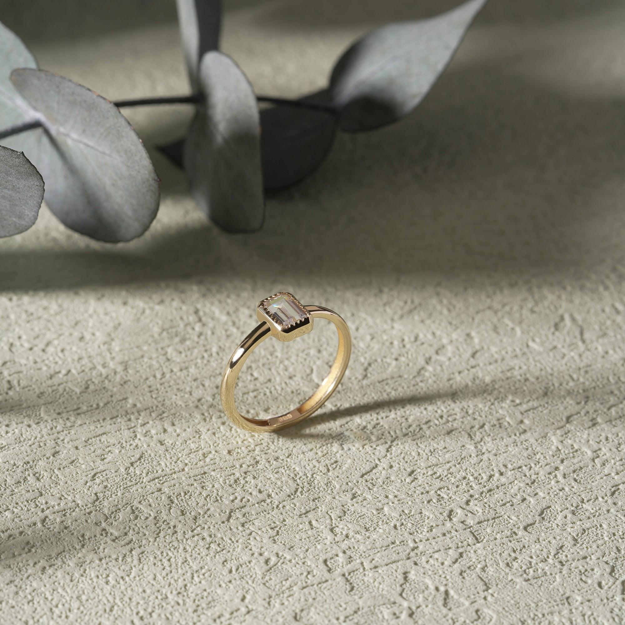 Gold ring with rectangular gemstone on textured surface, eucalyptus leaves in background