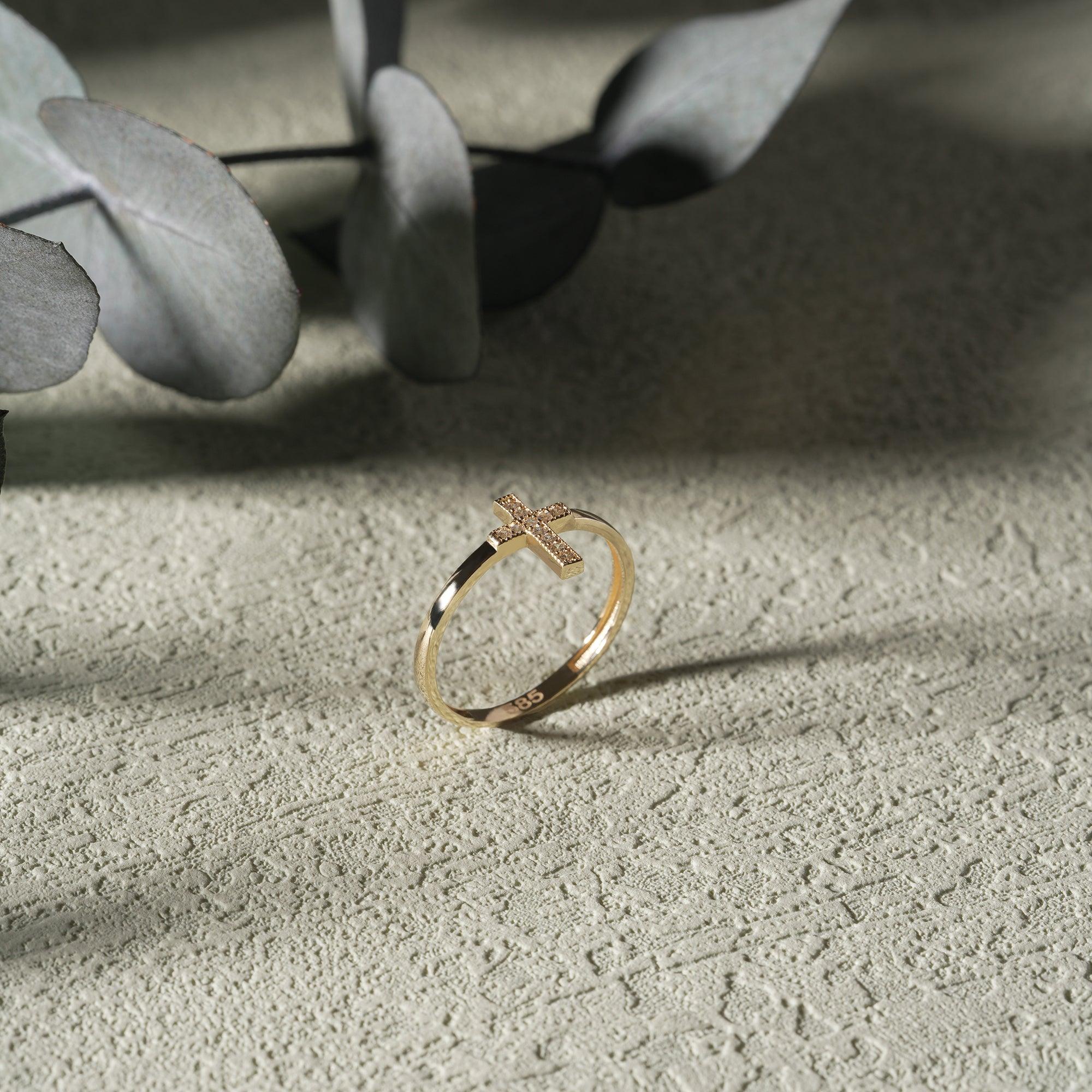 Gold cross ring with pavé stones on textured surface, minimalist jewelry photography.