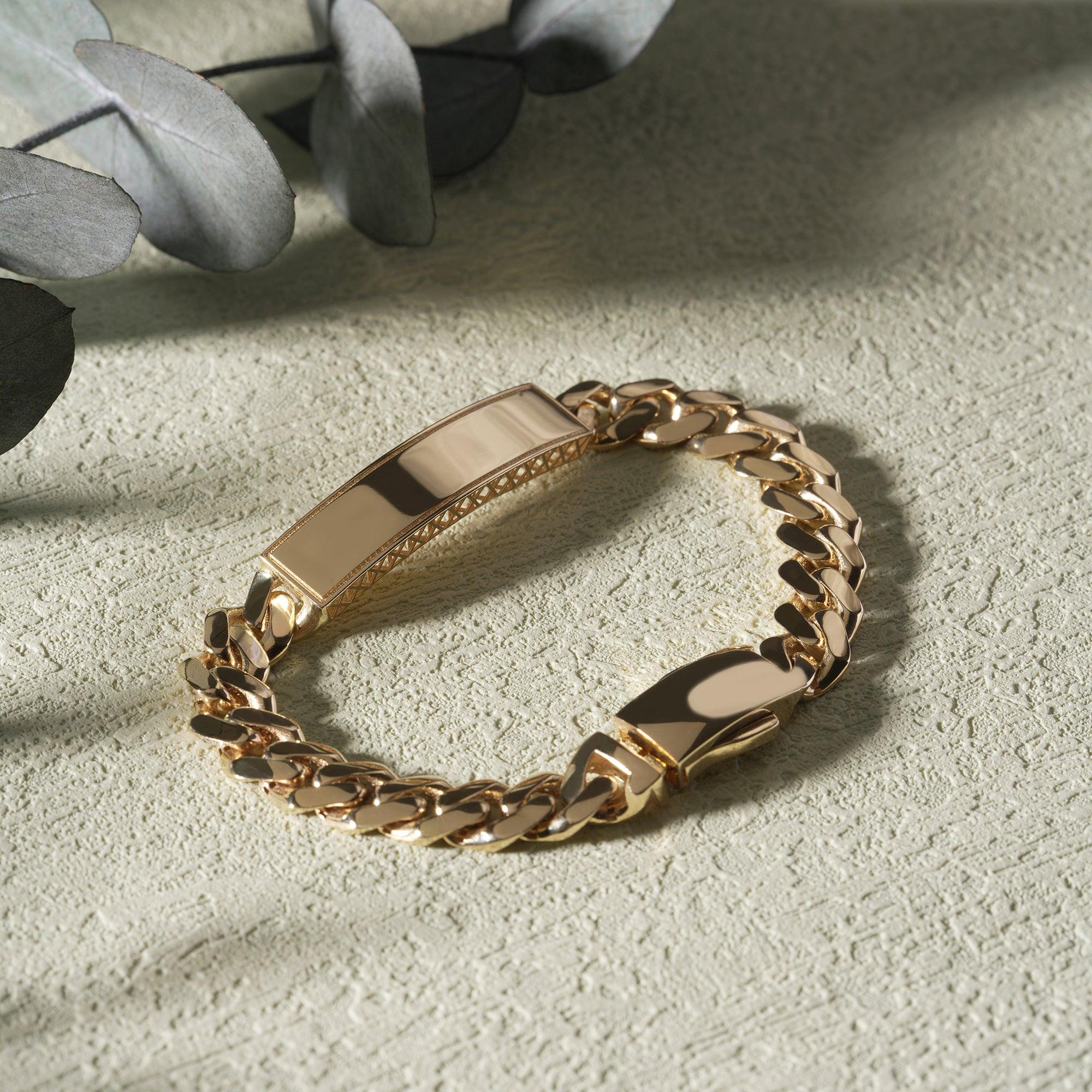 Polished gold ID chain bracelet on textured beige surface with leaves in background