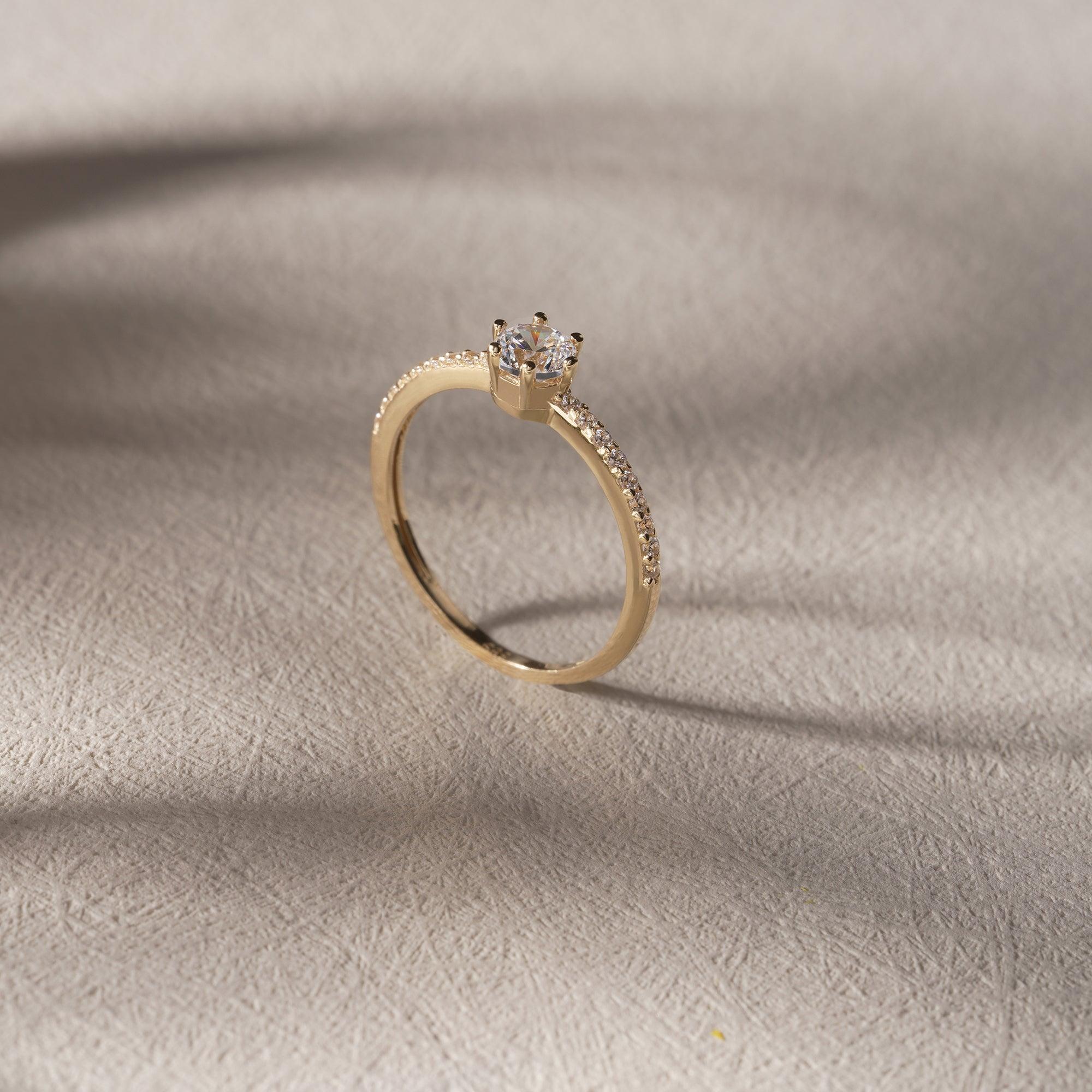 Gold solitaire engagement ring with pavé diamond band on textured beige surface