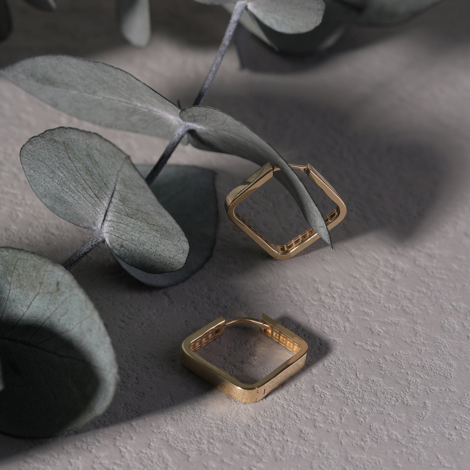 Gold square-shaped earrings on a textured surface with eucalyptus leaves.