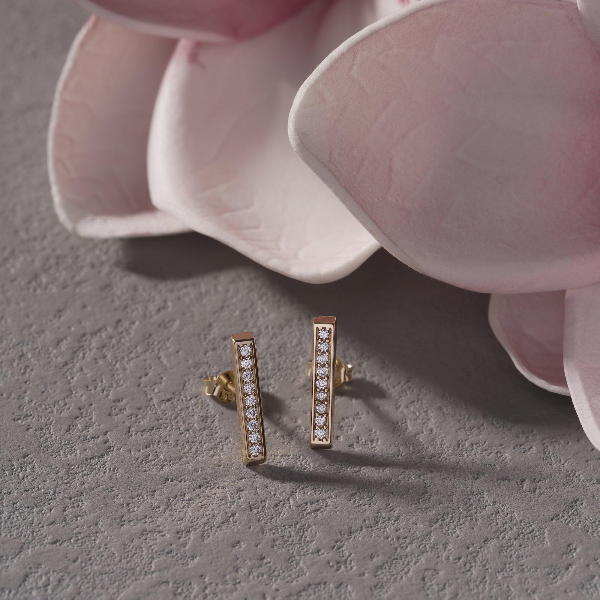 Gold bar stud earrings with diamonds on textured surface beside pink flower petals