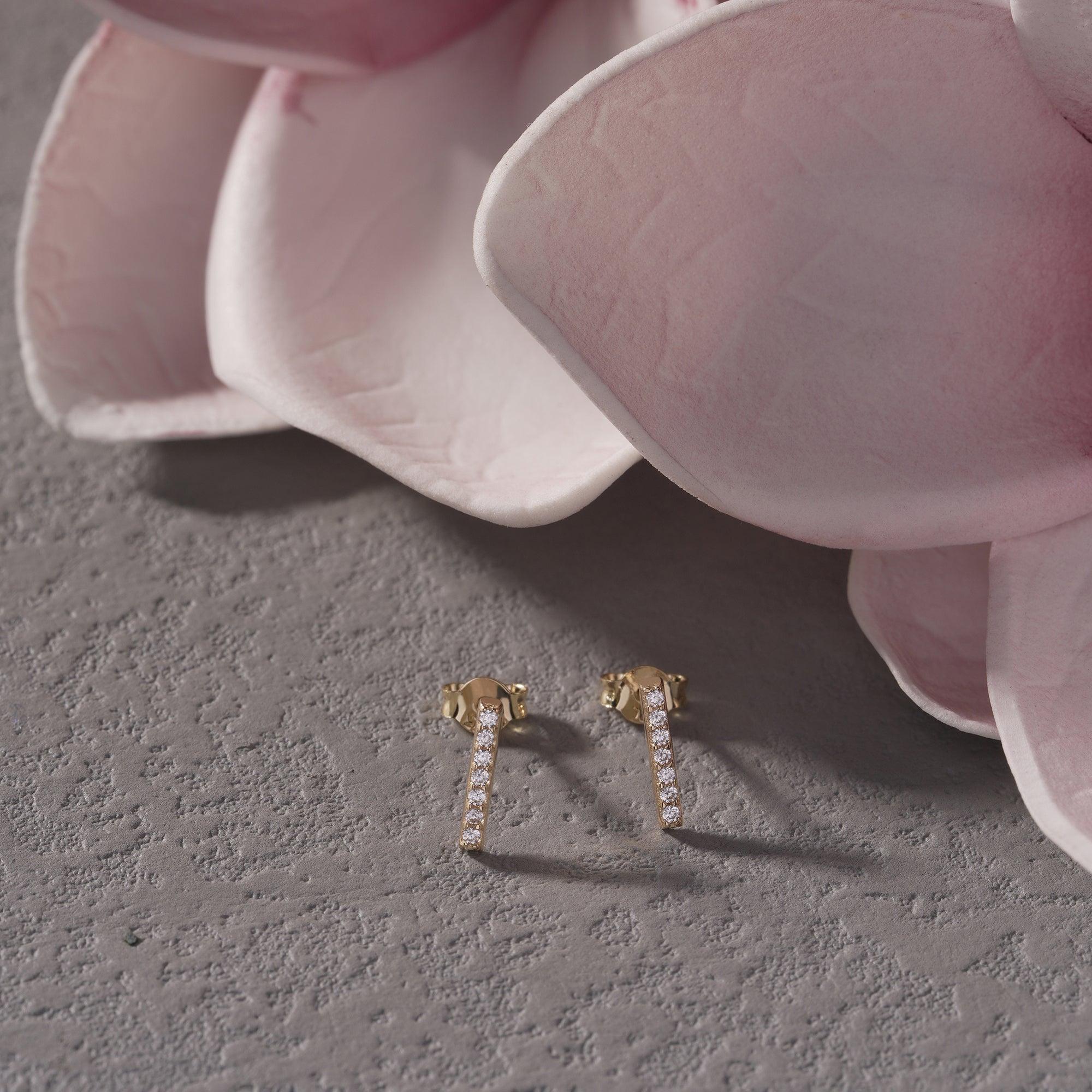 Gold bar stud earrings with diamonds on a textured surface beside pink flower petals