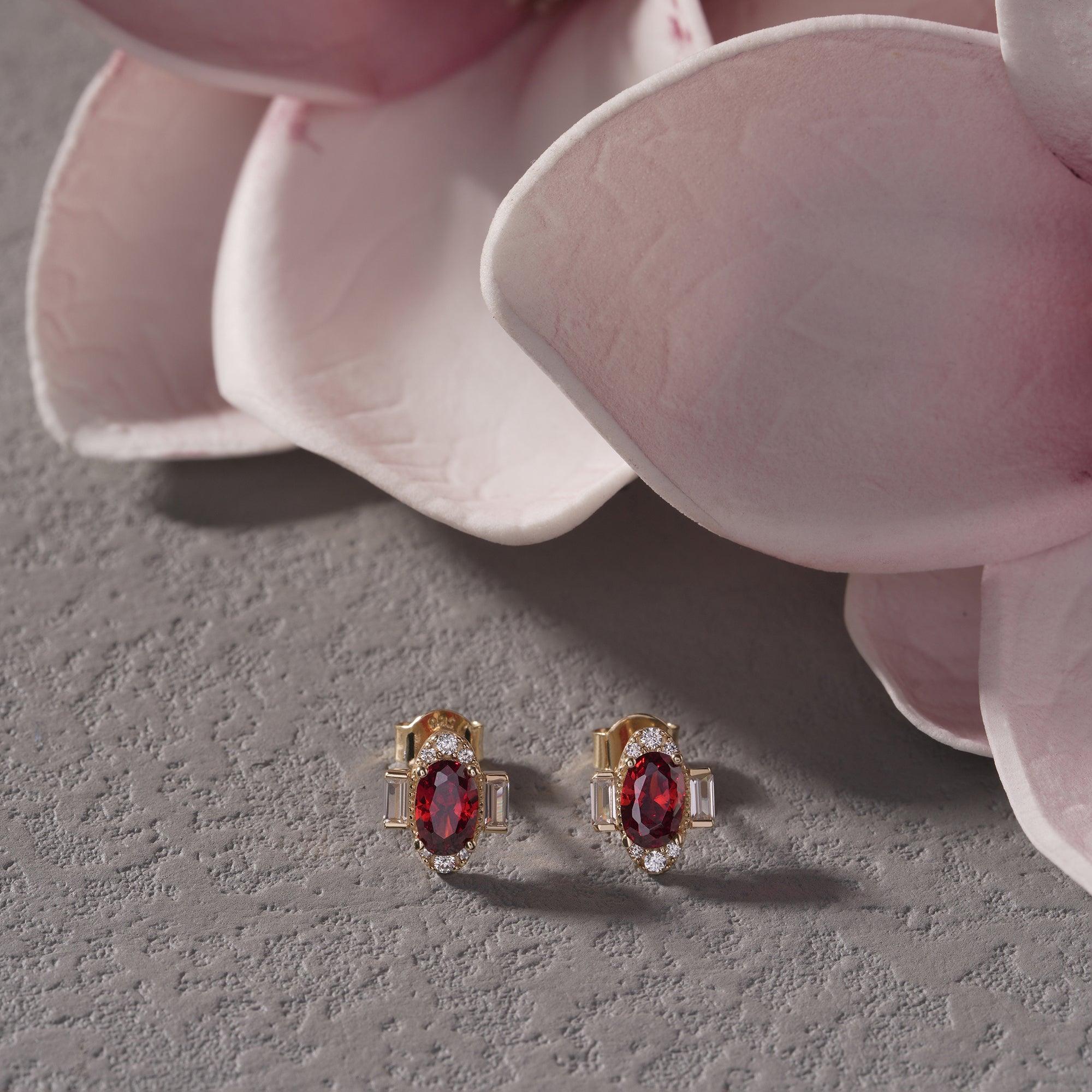 Gold earrings with oval red gemstones and clear accents on textured gray surface with pink flowers