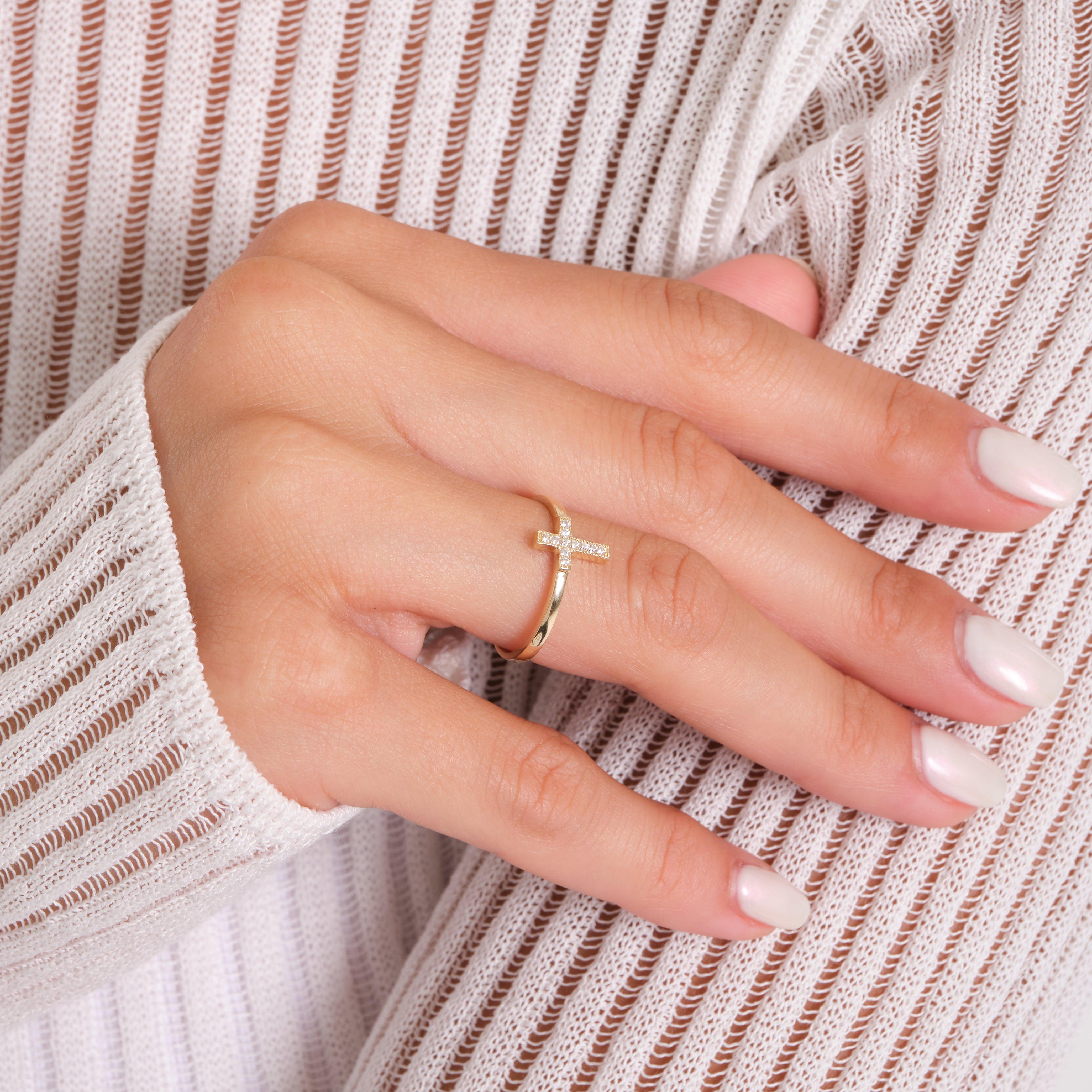 Gold cross ring with small diamonds on a woman's hand, wearing a white ribbed sleeve.