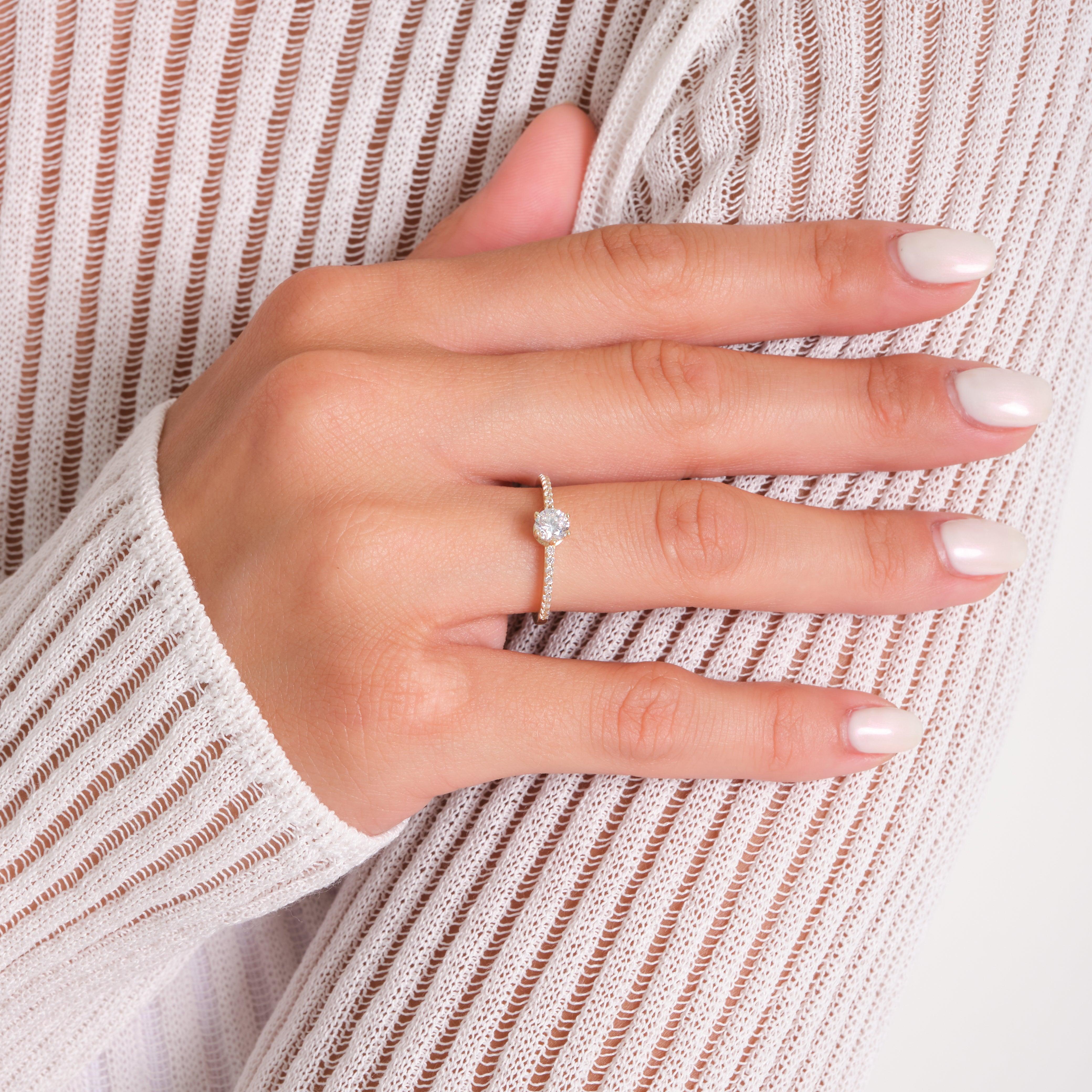 Woman’s hand wearing a gold diamond engagement ring with pavé band on neutral background