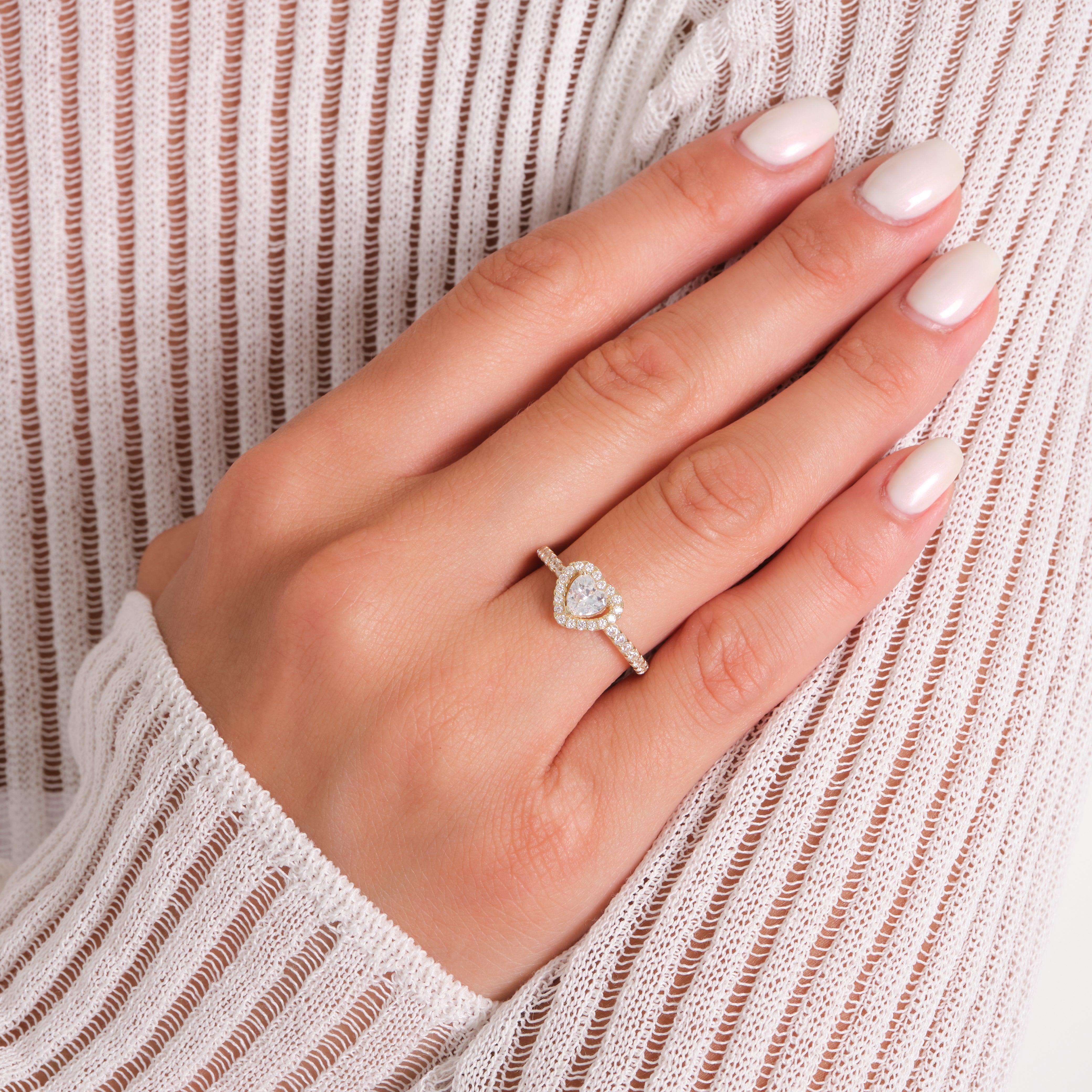 Gold heart-shaped diamond ring on hand with white polished nails and striped sweater