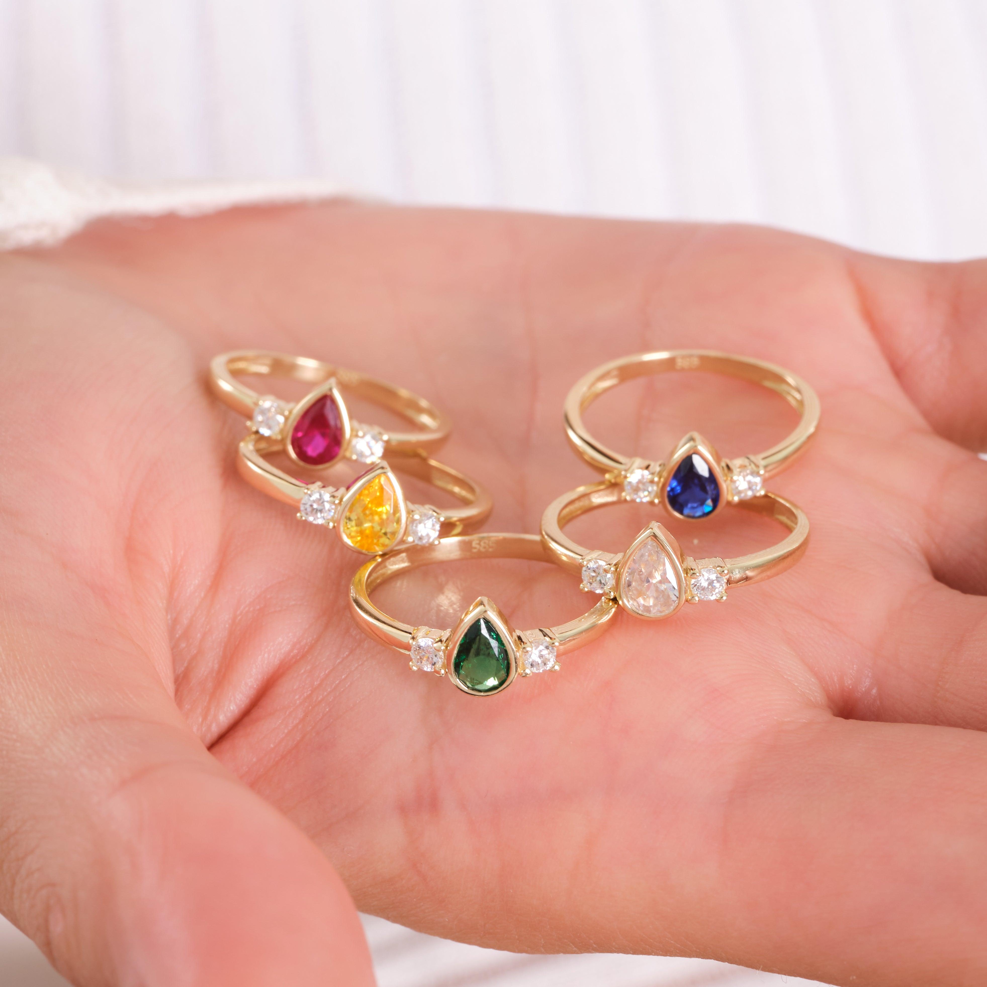 Hand holding gold rings with colorful teardrop gemstones and small diamond accents