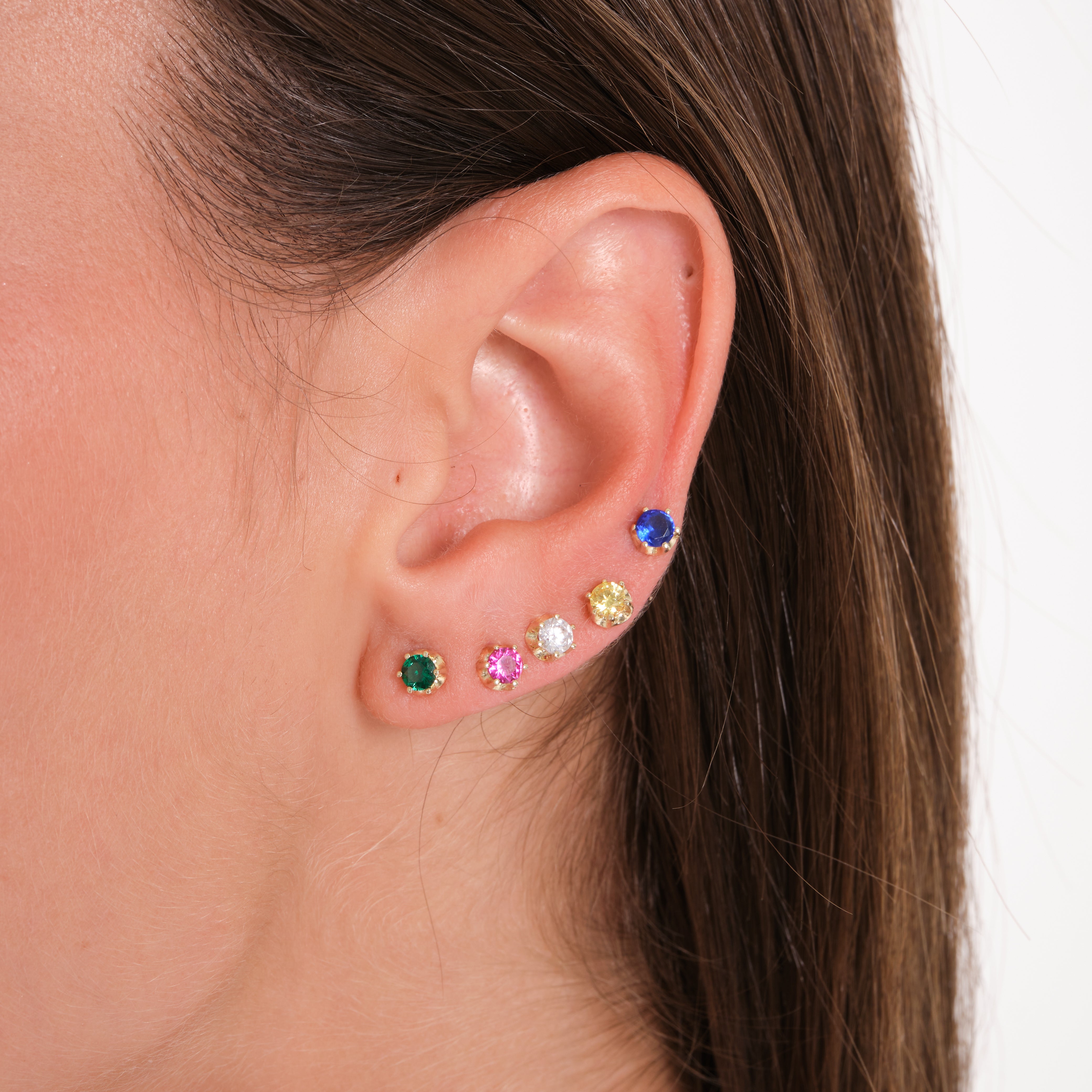 Close-up of an ear wearing colorful gemstone stud earrings with a neutral background.