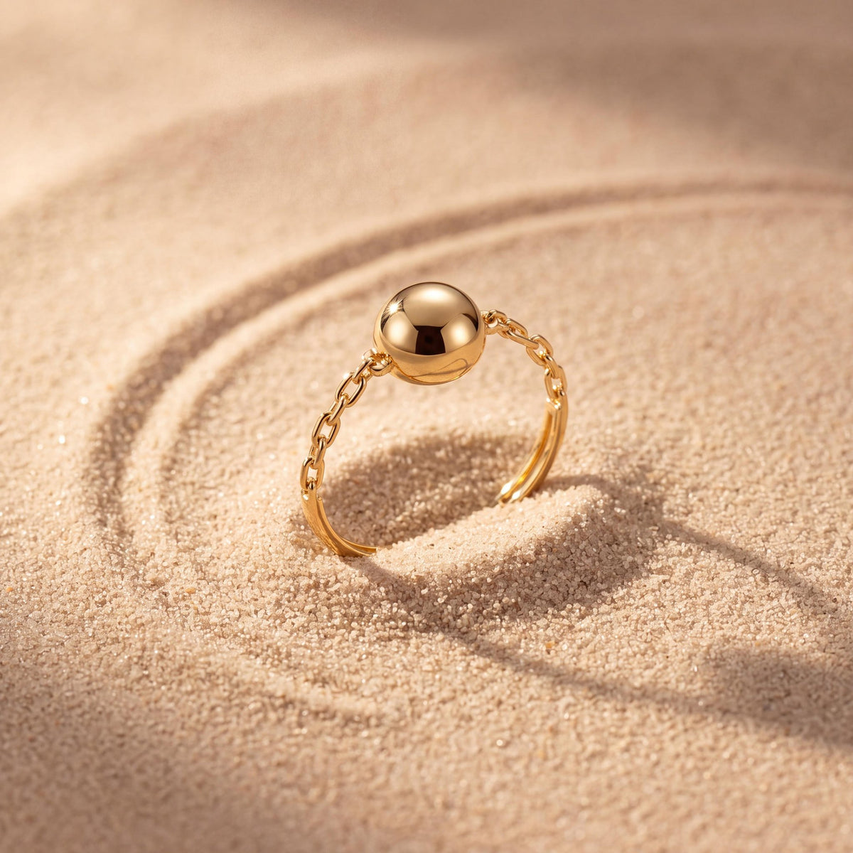 14K yellow gold ball ring standing upright in rippled sand with a long shadow
