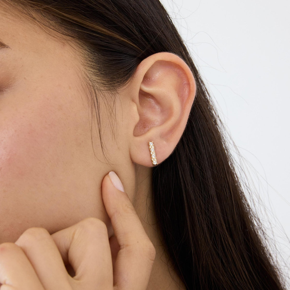 14K gold cubic zirconia bar stud earring worn in ear, model touching jawline with fingertip