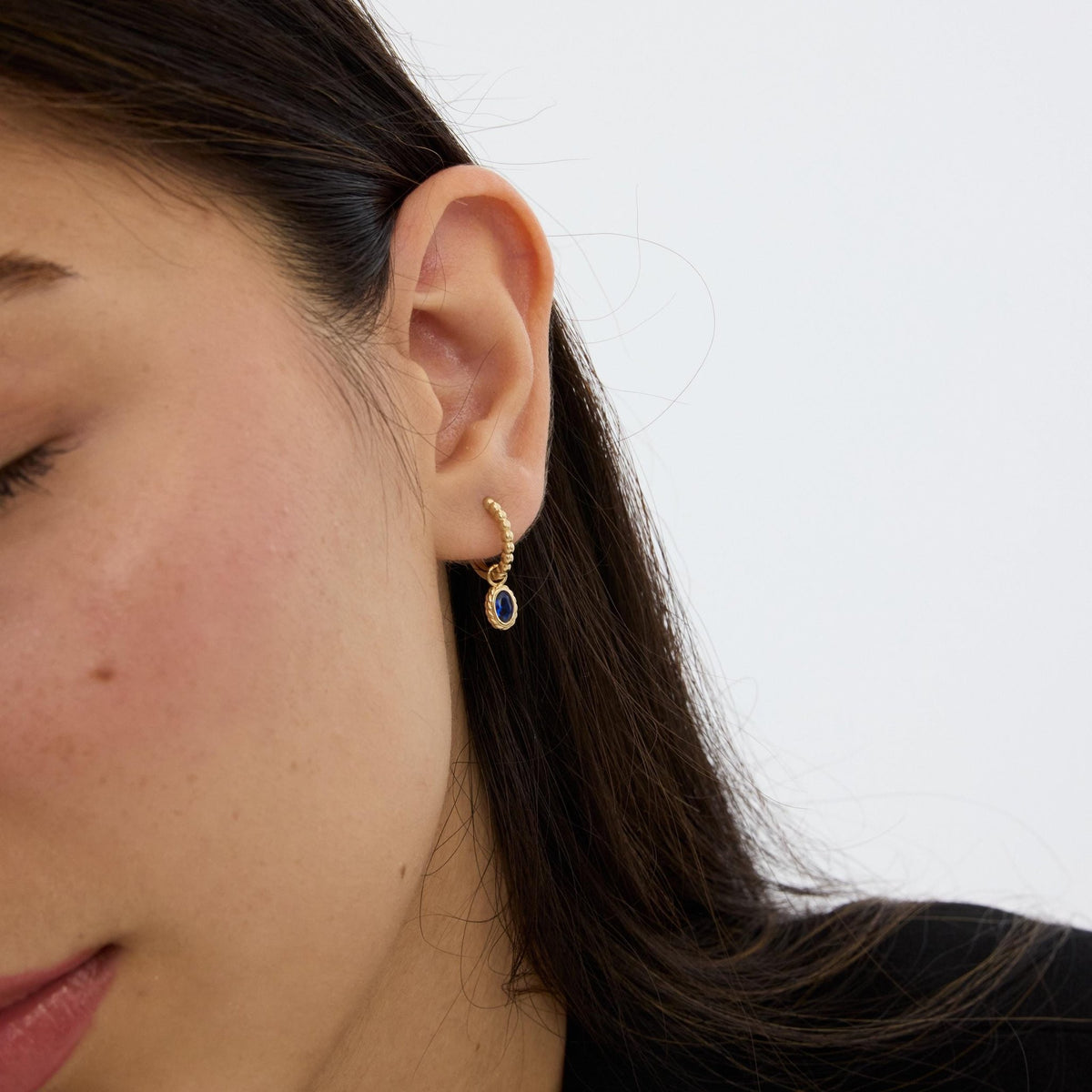 Woman's ear with 14K gold beaded huggie and dangling oval blue stone, face partly in frame