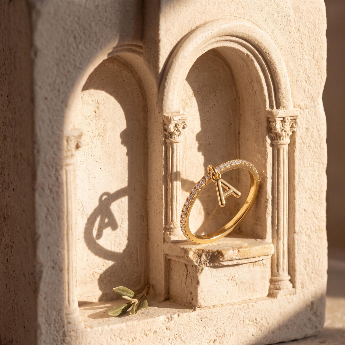 14K yellow gold dangle initial ring standing in a carved sandstone arch niche with an olive sprig