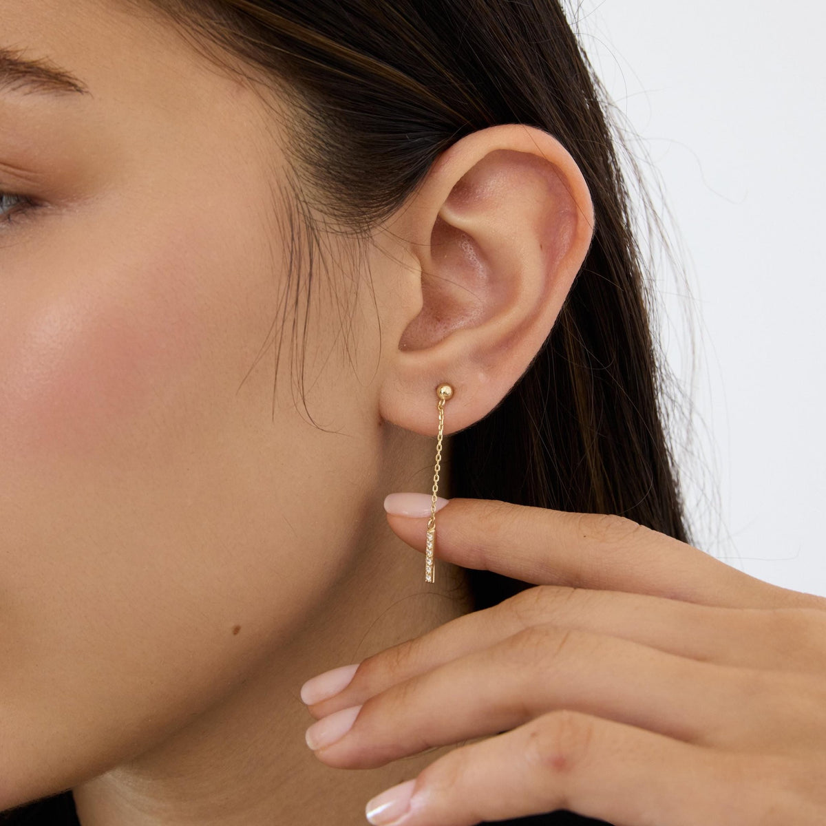 14K gold drop stud earring with round cubic zirconia bar worn on ear, hand touching the chain