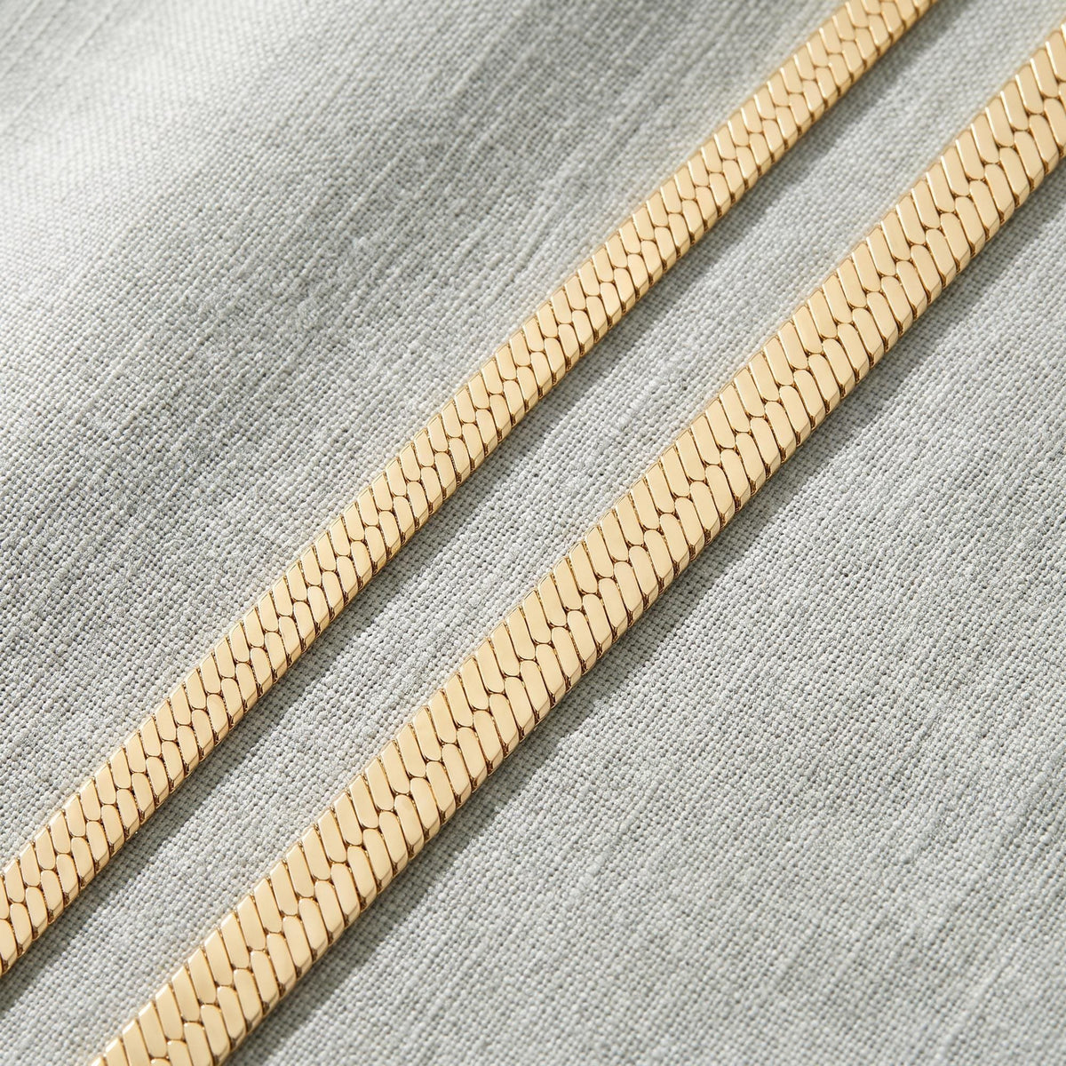 Close-up of two 14K yellow gold herringbone chains lying flat diagonally on grey linen fabric