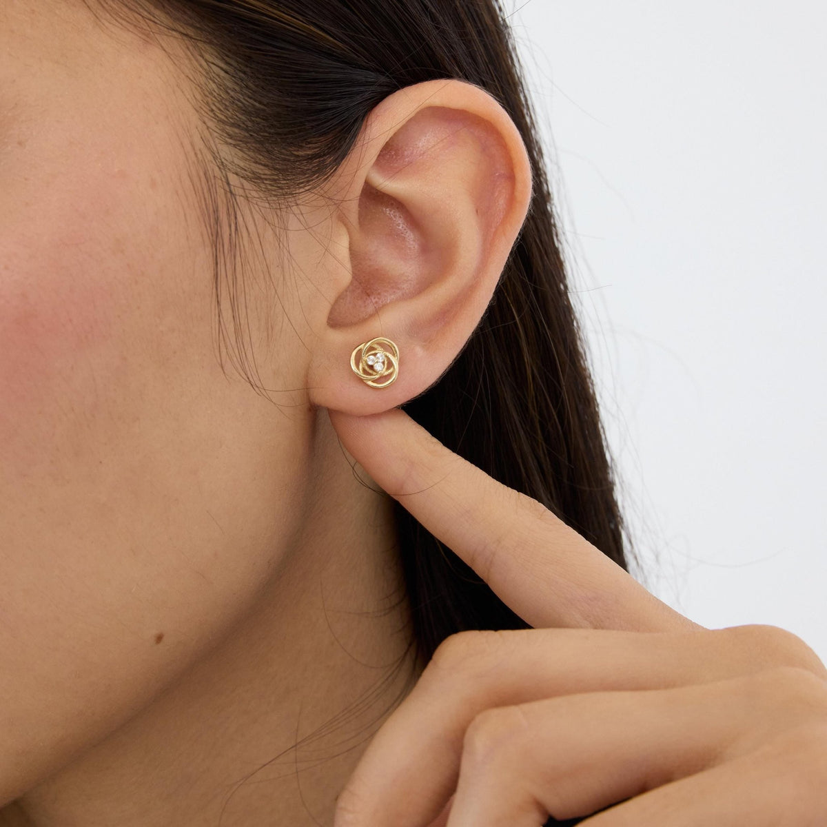 14K yellow gold love knot stud earring with cubic zirconia on ear, close crop with fingertip under earlobe