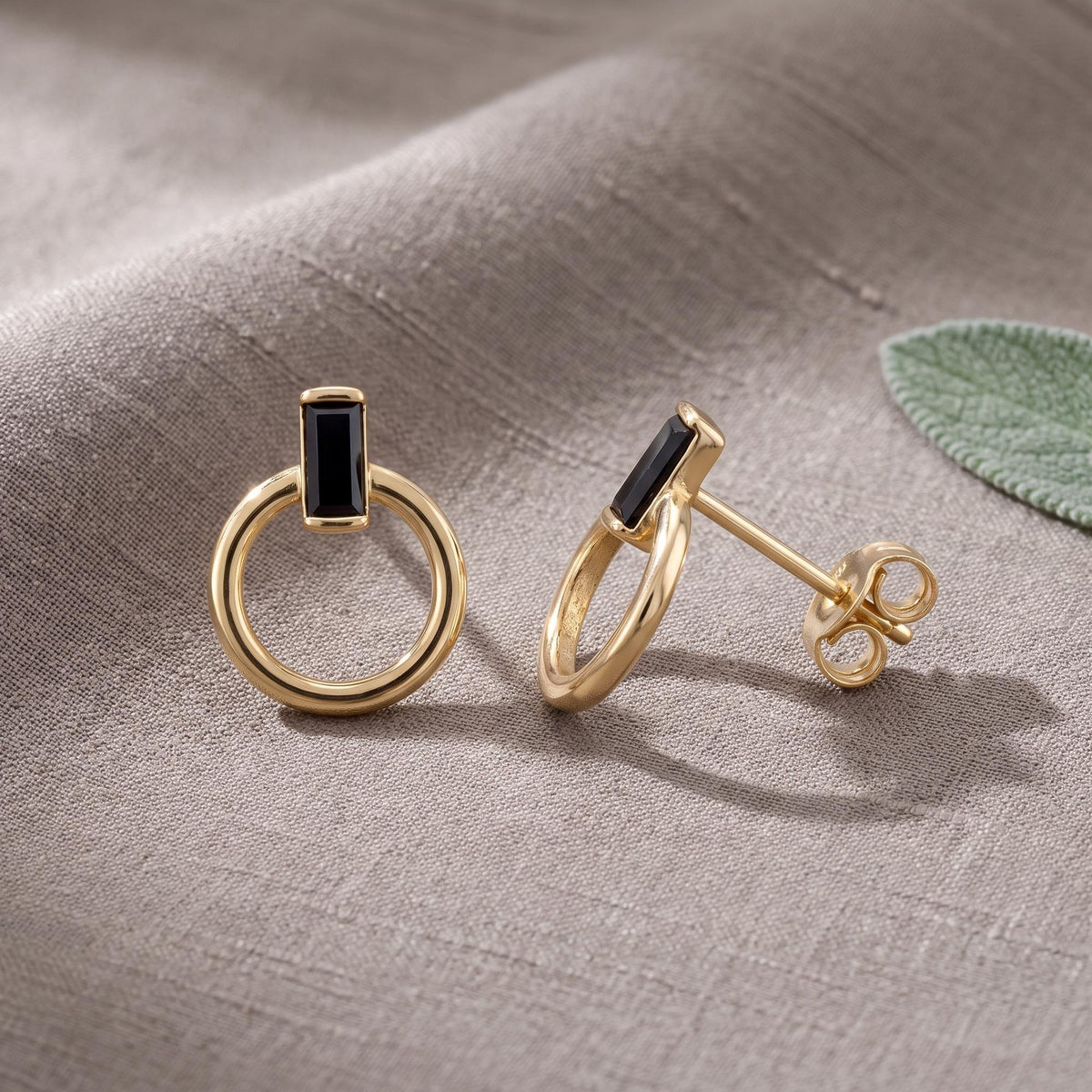 14K yellow gold open circle stud earrings with black baguette stone, pair resting on grey linen beside a leaf