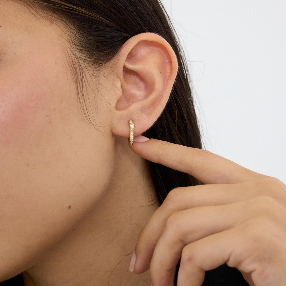 Woman touching her ear to show a 14K gold square huggie hoop earring with cubic zirconia