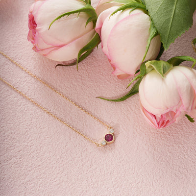 Gold necklace with a red gemstone pendant on a pink surface with pink roses.