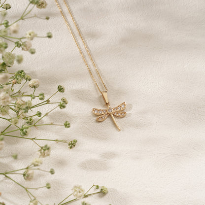 Gold necklace with a dragonfly pendant on a soft beige background with delicate flowers.