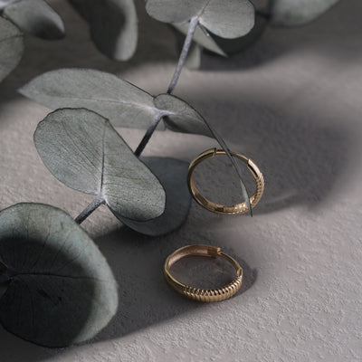 Two gold hoop earrings on a gray surface with eucalyptus leaves.