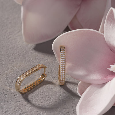 Gold hoop earrings with pavé diamonds on a stone surface beside pink flowers