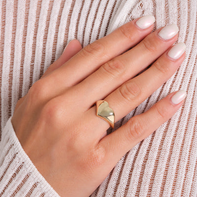 Hand wearing a gold heart-shaped ring with light nail polish on a textured fabric background