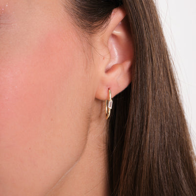 Close-up of an ear wearing a gold hoop earring with a clear gemstone on a neutral background