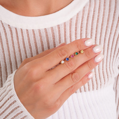 Hand wearing gold rings with colorful gemstone settings on index finger, white ribbed top