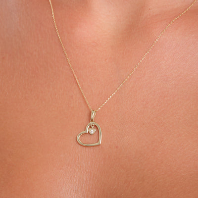 Gold heart-shaped necklace with a pearl pendant on a person's neck.