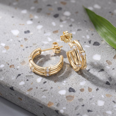 14K gold bamboo hoop earrings with round cubic zirconia on a speckled stone surface beside a green leaf