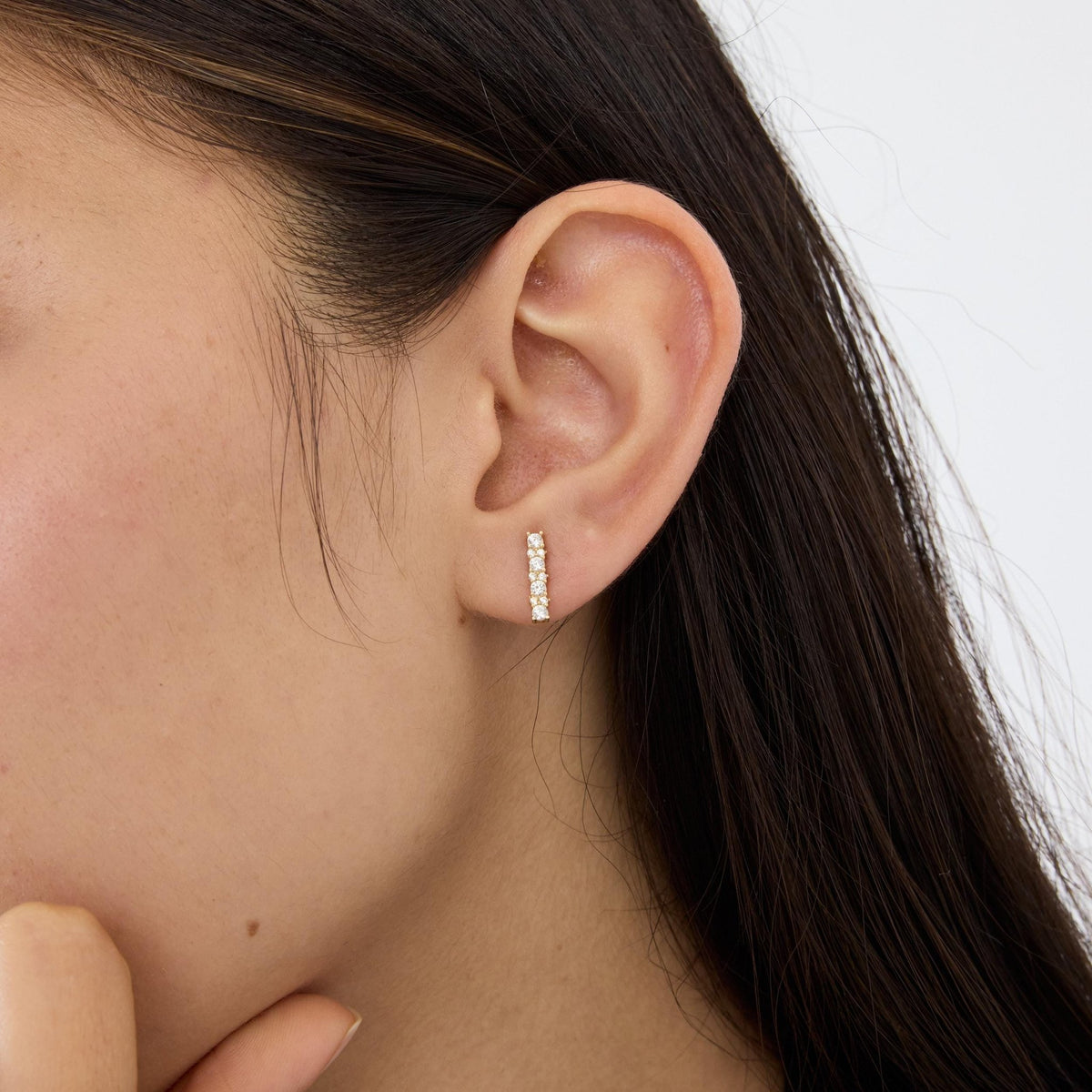 14K gold cubic zirconia bar stud earring worn in earlobe, close-up with fingers at bottom of frame